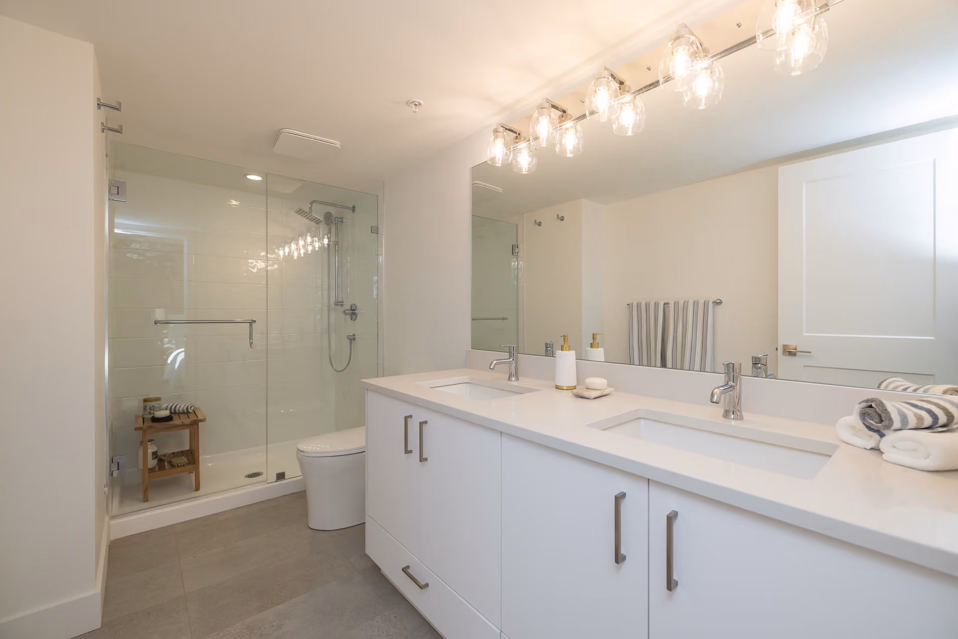 Skyview – A modern, bright bathroom featuring a white double vanity with quartz countertops and sleek silver handles under a large mirrored vanity light fixture. A glass-enclosed walk-in shower with white tile and a small wooden bench sits next to a modern toilet against warm white walls and large grey floor tiles.