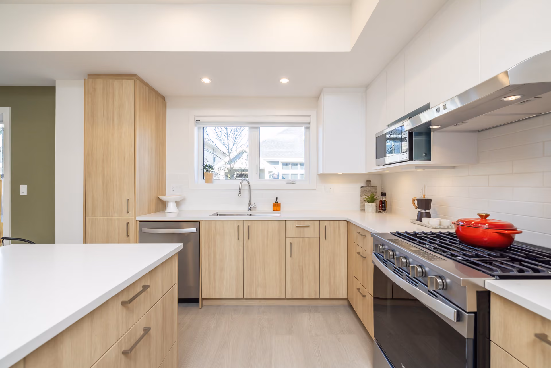 Skyview – A modern, bright kitchen featuring light wood lower cabinetry, flat-panel white upper cabinets, white quartz countertops, and a white subway tile backsplash. Stainless steel appliances include a gas range with a red Dutch oven, a range hood, an over-the-range microwave, and a dishwasher set under a window.