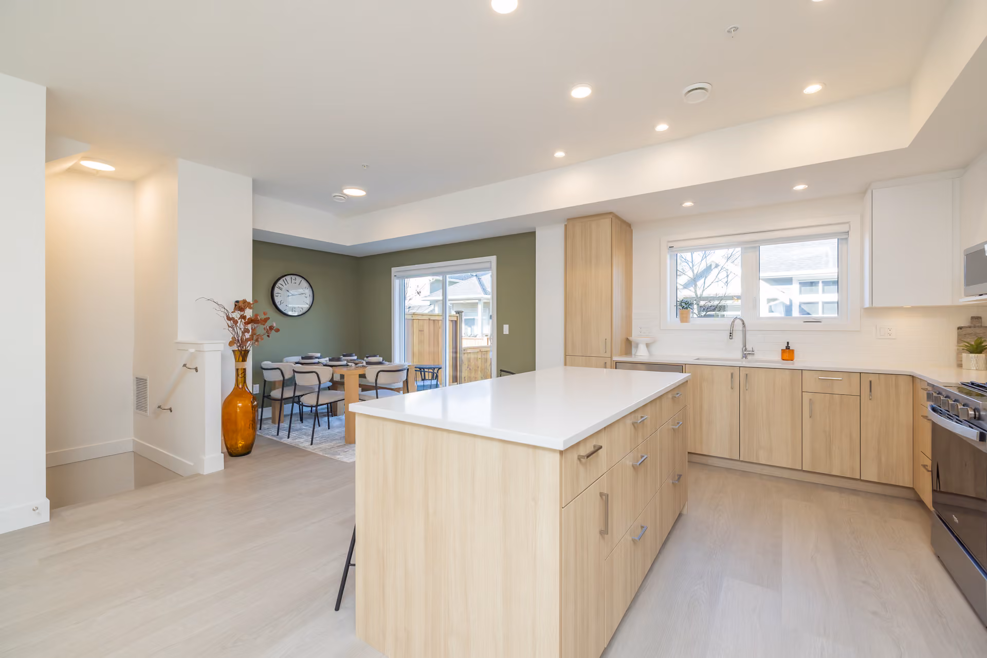Skyview – A bright, open-concept kitchen and dining area featuring light wood cabinetry, white quartz countertops, and a matching kitchen island. In the background, a dining table sits against an olive green accent wall next to glass sliding doors that open to an outdoor patio.