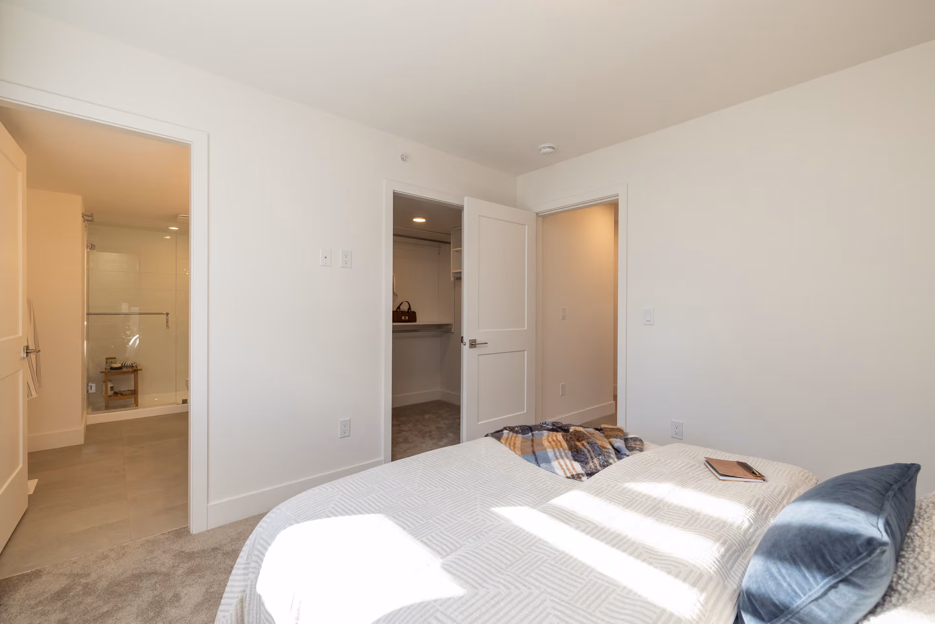 Skyview – A modern bedroom featuring plush grey carpeting, a bed dressed in light-patterned linens, and open doorways leading to a walk-in closet and an en-suite bathroom with a glass-enclosed shower. Bright natural light streams across the bed and off-white walls, creating a clean, inviting atmosphere.