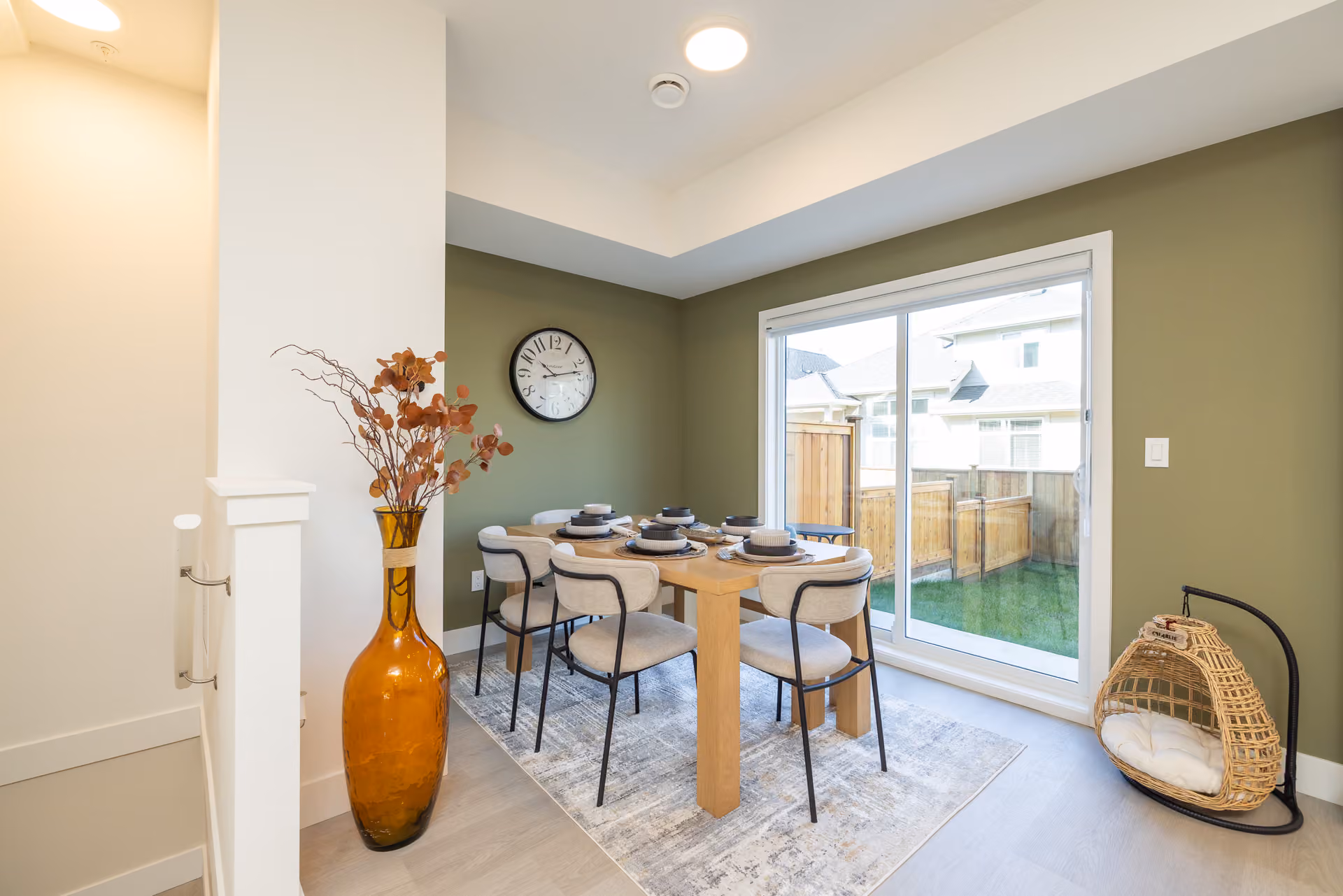 Skyview – A bright dining area features a wooden table set for six with upholstered chairs over an area rug, highlighted by olive green accent walls and a large wall clock. A sliding glass door leads out to a fenced backyard patio, while a large amber glass floor vase sits in the foreground.