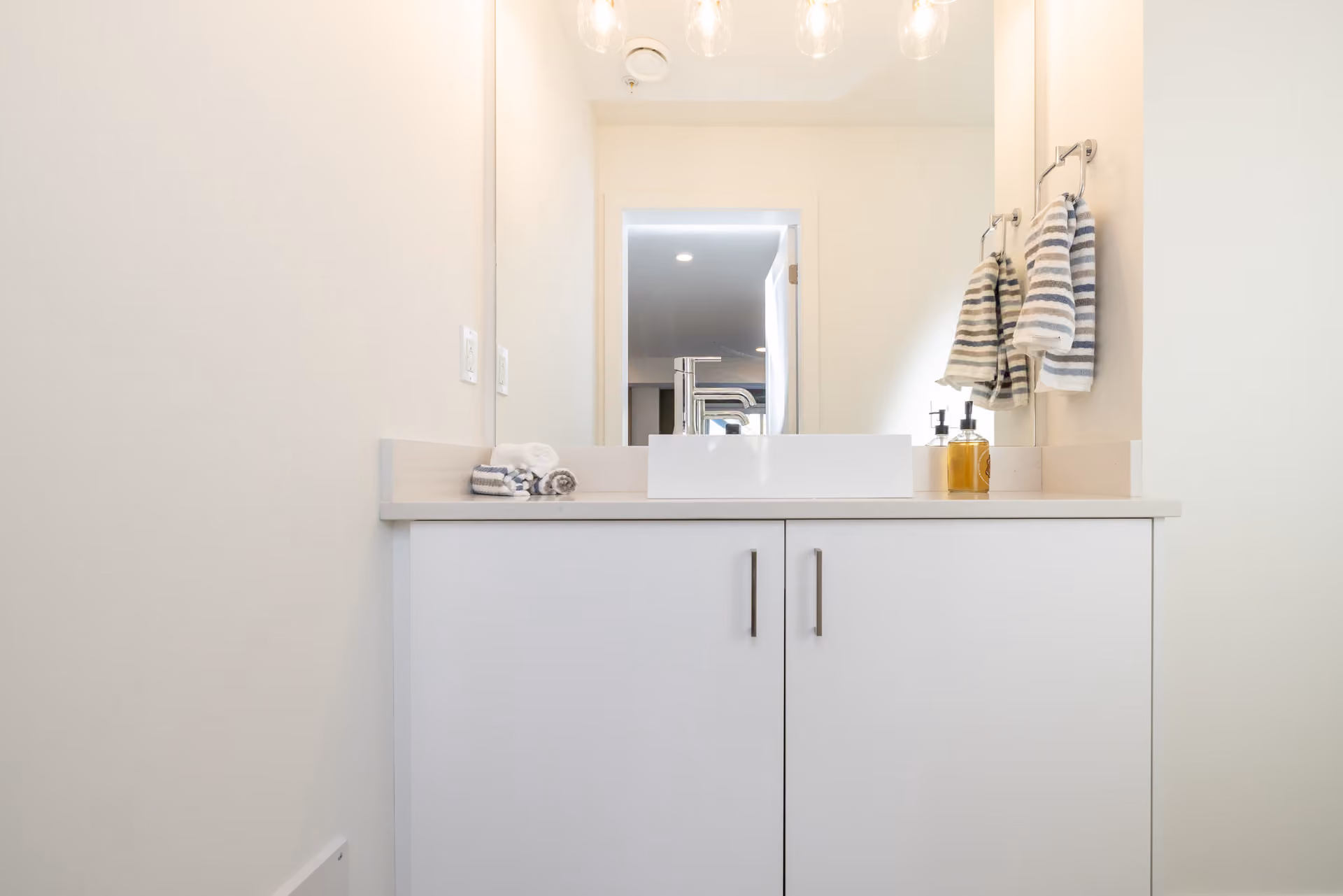 Skyview – A clean, modern bathroom vanity featuring a white vessel sink atop a light beige countertop and minimalist white cabinetry. Above the vanity, a large wall-to-wall mirror reflects a doorway and pendant light fixtures under bright, neutral interior lighting.