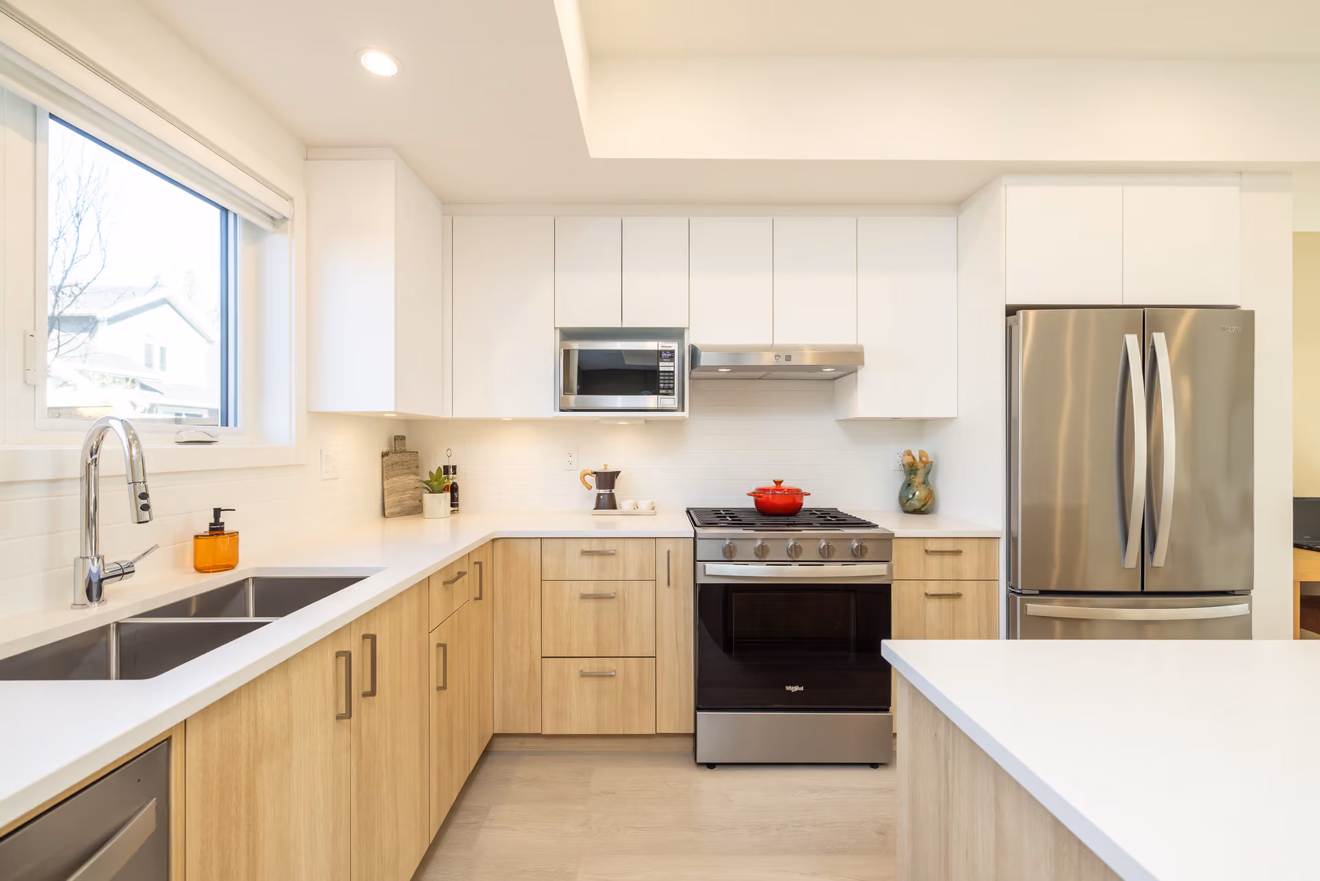 Skyview – A modern, bright L-shaped kitchen featuring two-tone cabinetry with light natural wood lower cabinets and flat-panel white upper cabinets. Stainless steel appliances include a gas range with a red Dutch oven on top, an over-the-range microwave, and a French door refrigerator set against white quartz countertops.