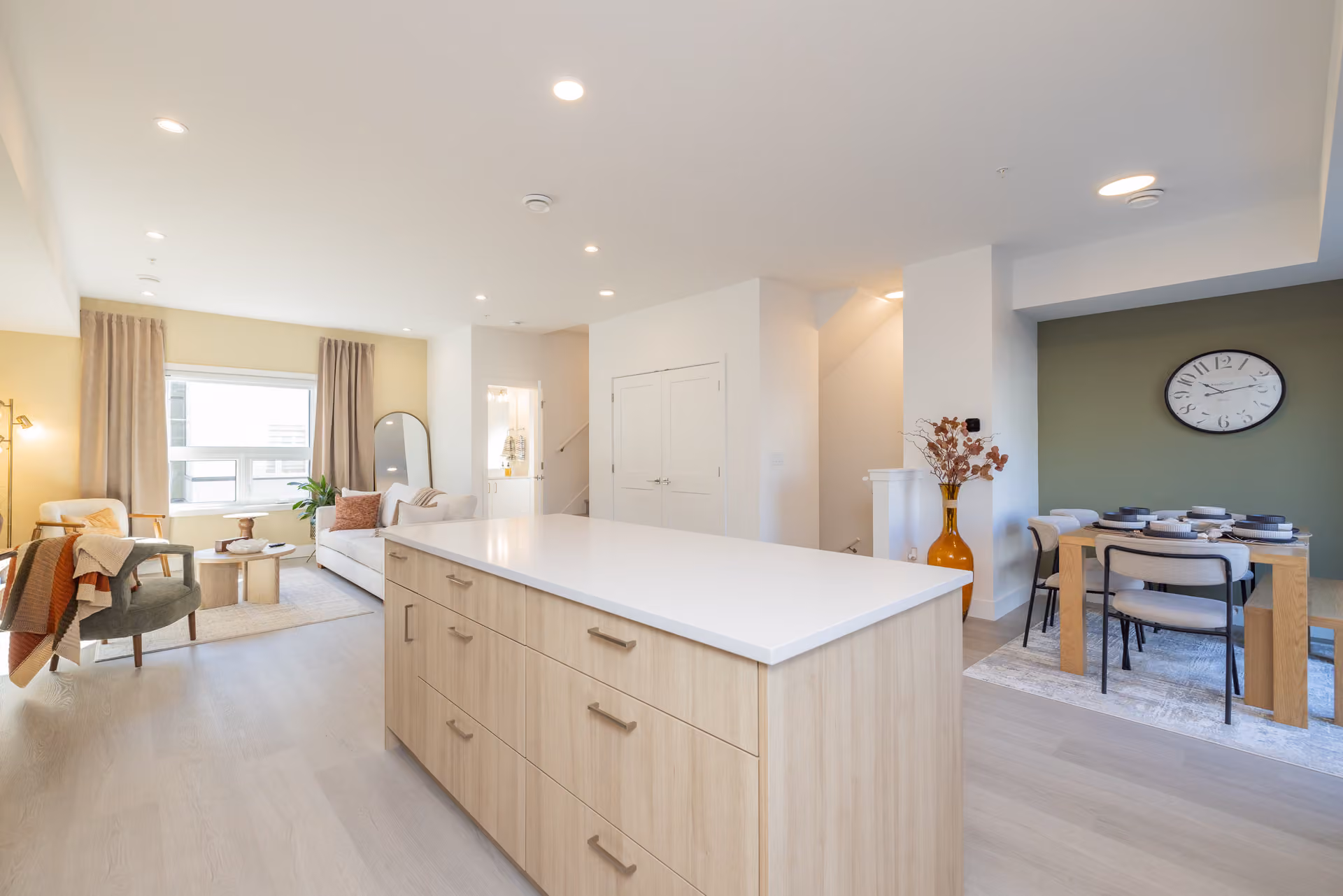 Skyview – A bright, open-concept interior featuring a light-wood kitchen island with a solid white countertop in the foreground. In the background, the space seamlessly flows into a living room with comfortable seating and a dining nook highlighted by an olive-green accent wall and large wall clock.
