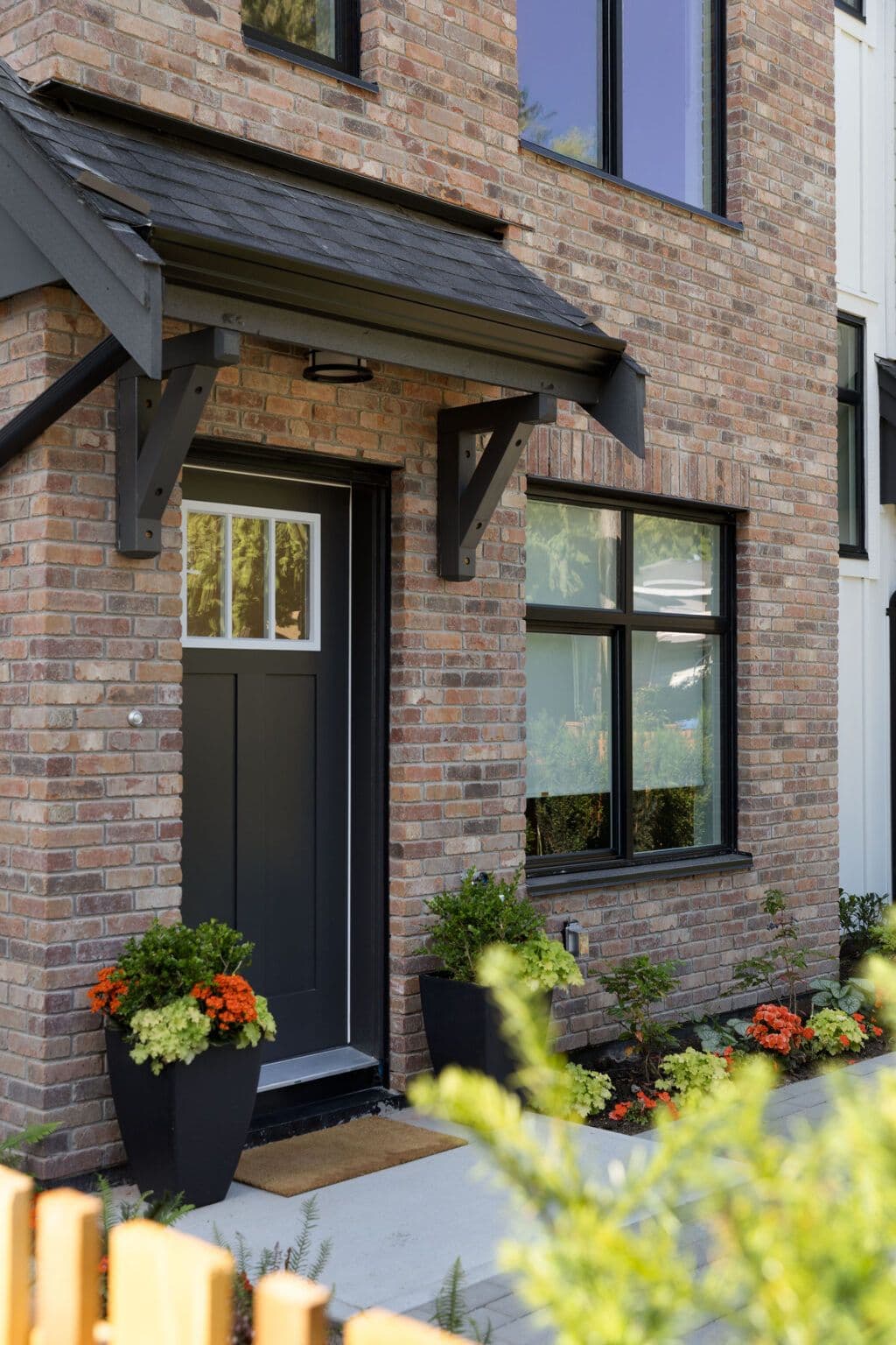 Rindall – Close-up of the brick building entrance with a black front door, covered awning, and potted flowering plants.