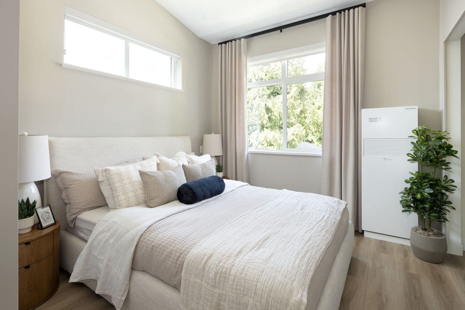 Rindall – Bedroom with a beige upholstered bed, wood nightstand, and a large white indoor heat pump unit beside a plant.