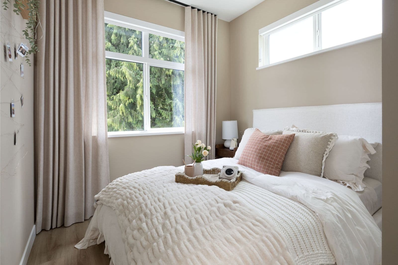 Rindall – Bedroom with an upholstered bed, string-light photo display on the wall, and a large window looking out to trees.