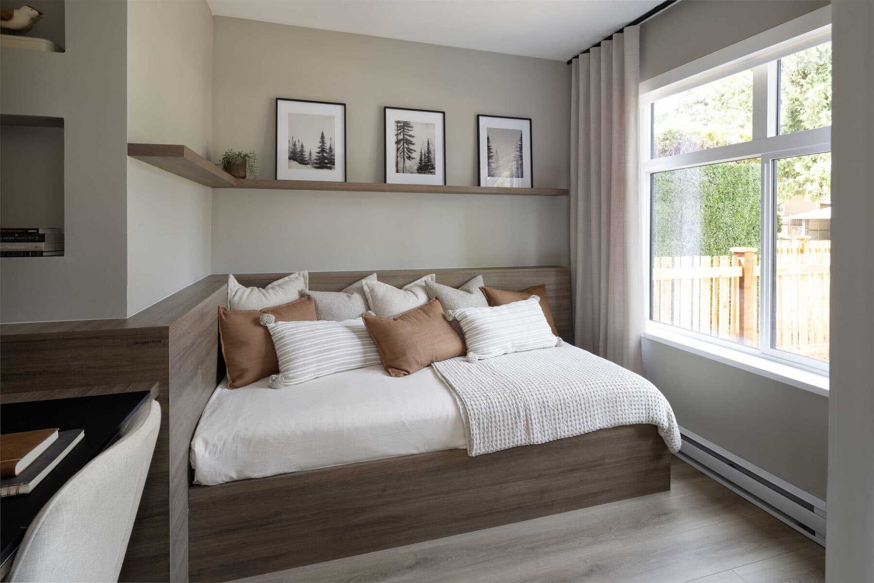 Rindall – Bedroom nook with a built-in daybed, floating wood shelf with framed prints, and a window looking onto a fenced yard.