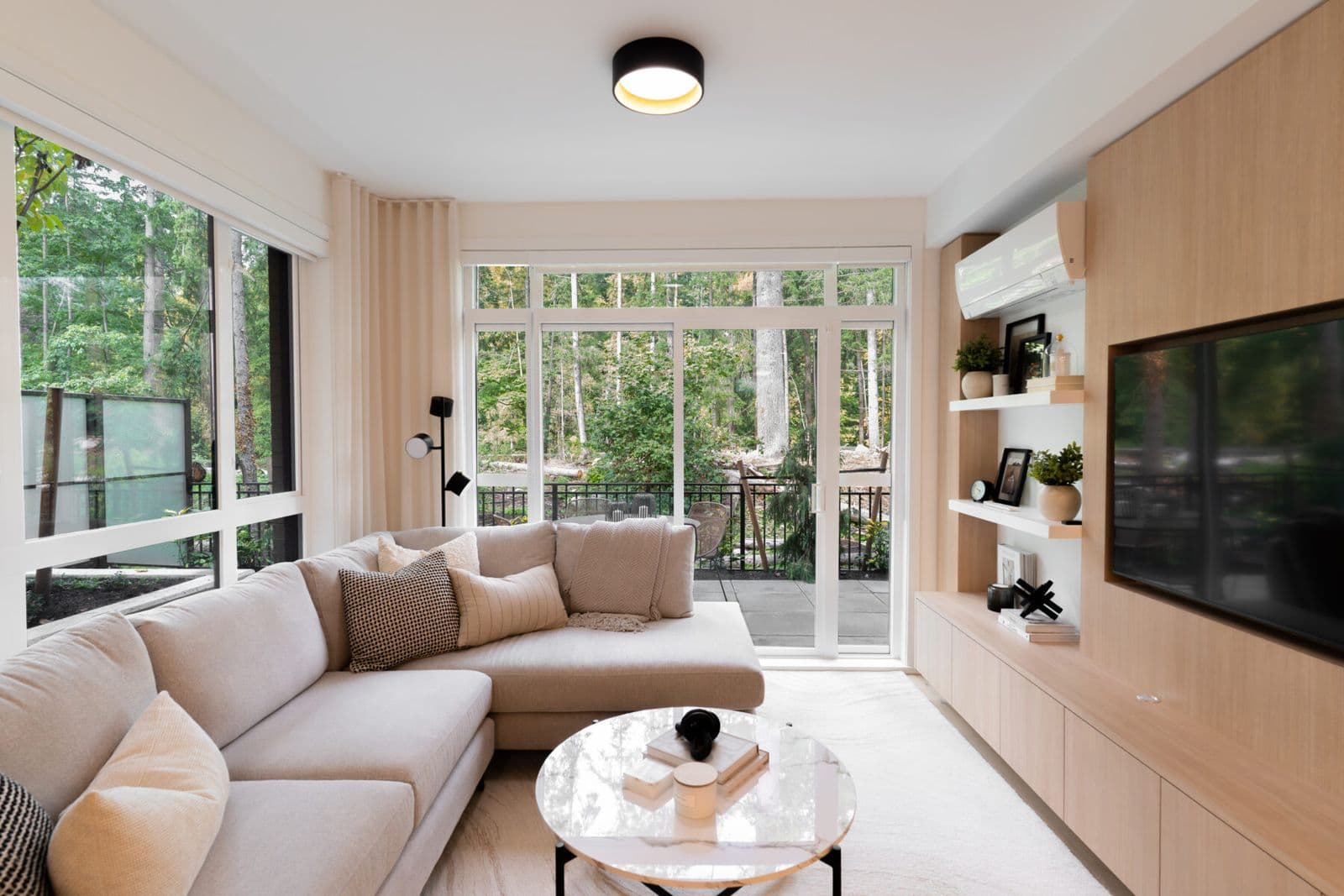Rindall – Living room with a beige sectional, wood-paneled TV wall, and sliding glass door opening onto a wooded balcony view.