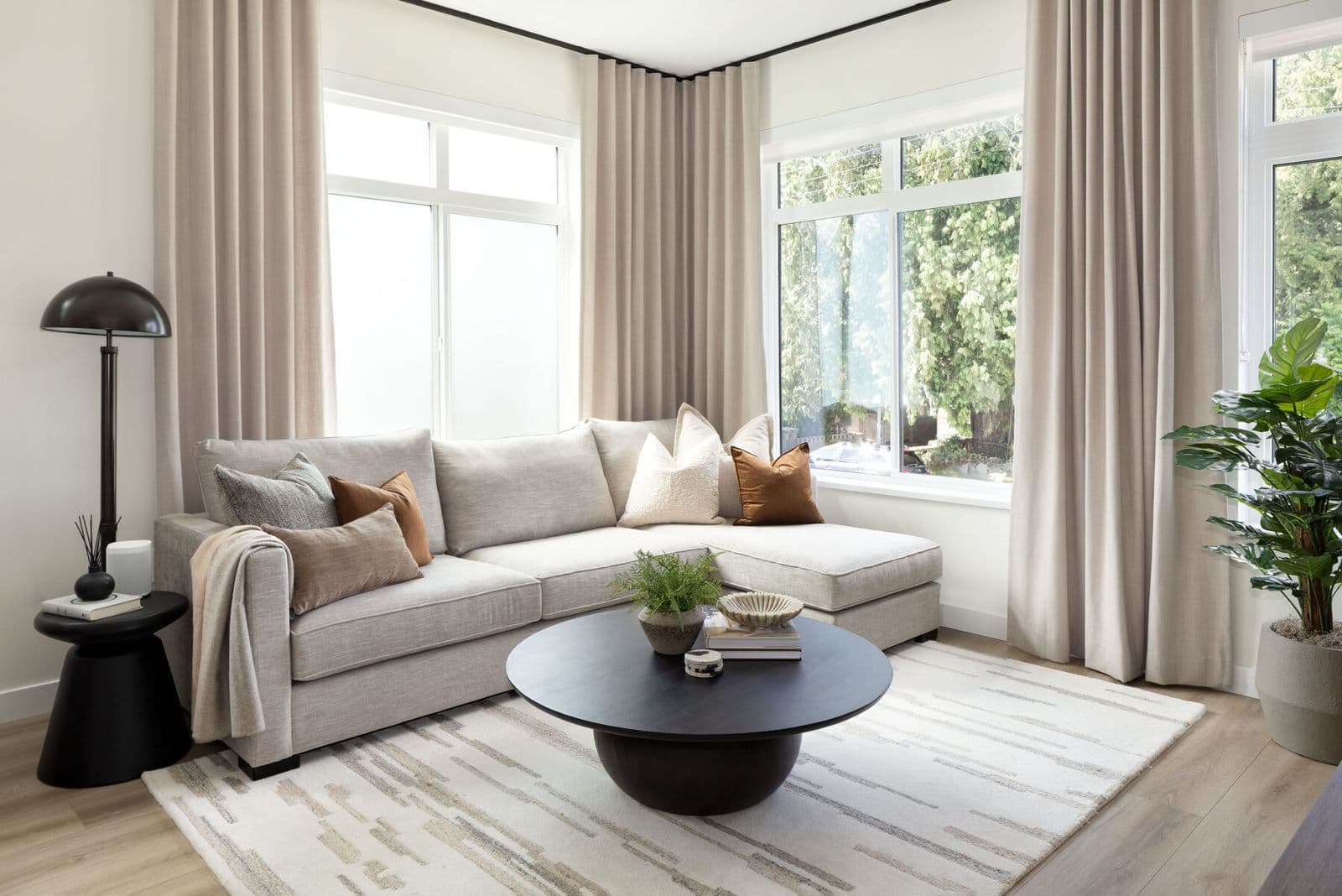 Rindall – Living room corner with a beige sectional sofa, dark round coffee table, and large windows with sheer curtains.