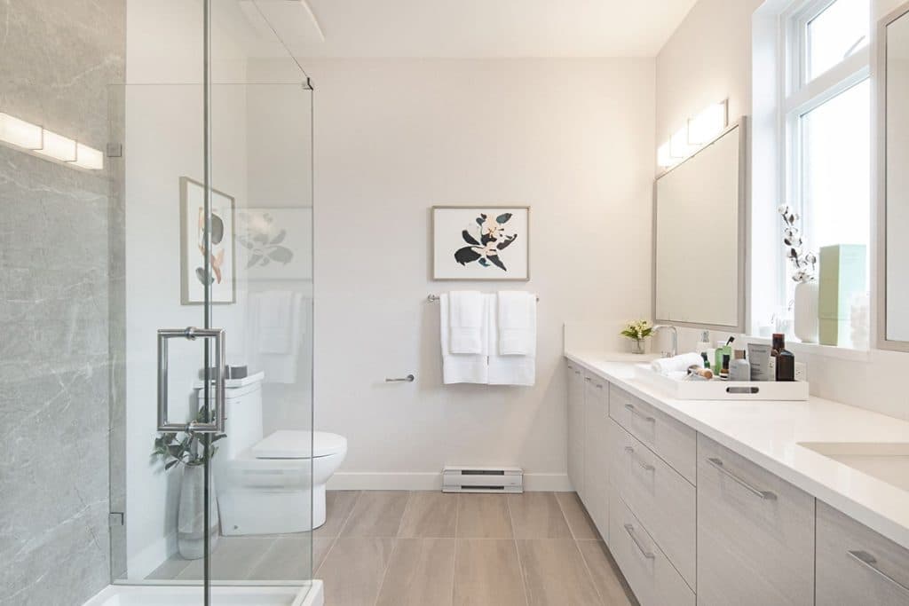 Baycrest West Condos – Bathroom with a long light-wood double vanity, framed floral art, towel bar, and a glass shower enclosure with stone tile.