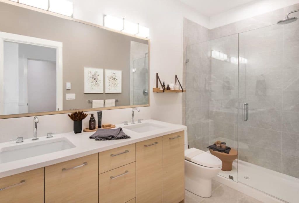 Baycrest West Condos – Bathroom with a wood double vanity, framed floral art, and a glass-enclosed walk-in shower next to the toilet.