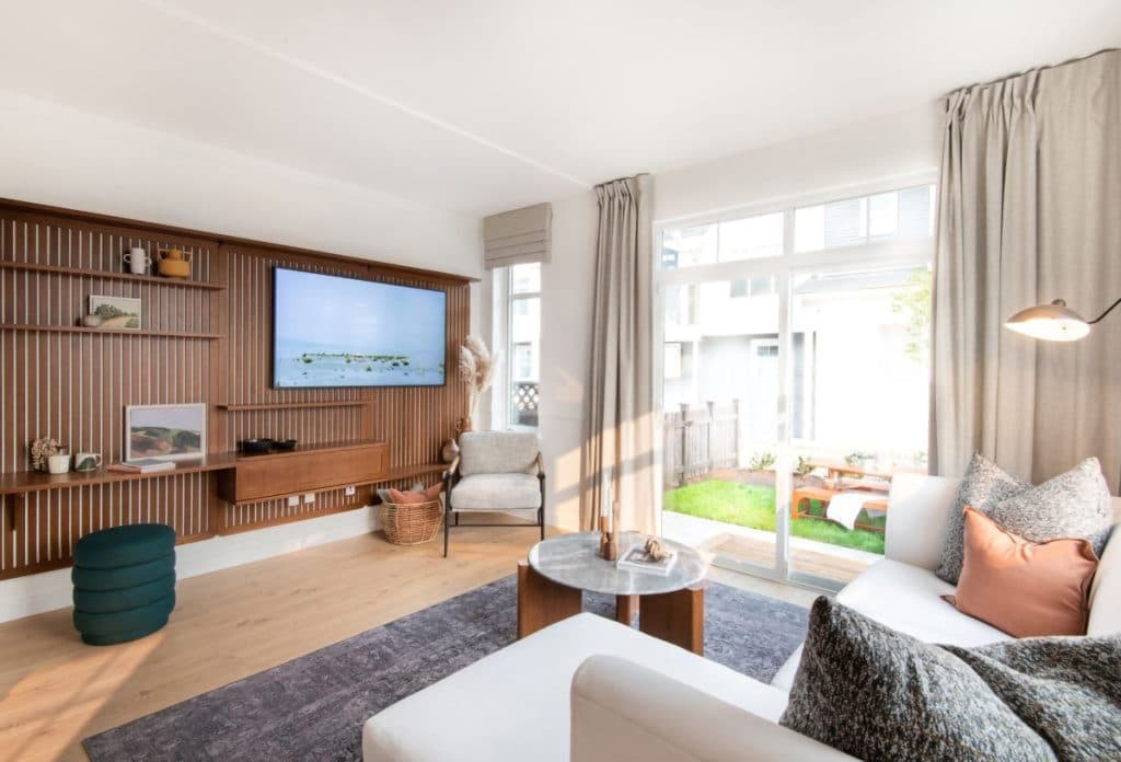 Baycrest West Condos – Living room with a slatted wood media wall and mounted TV, a white sofa with gray and terracotta pillows, and sliding glass doors opening to a fenced backyard.