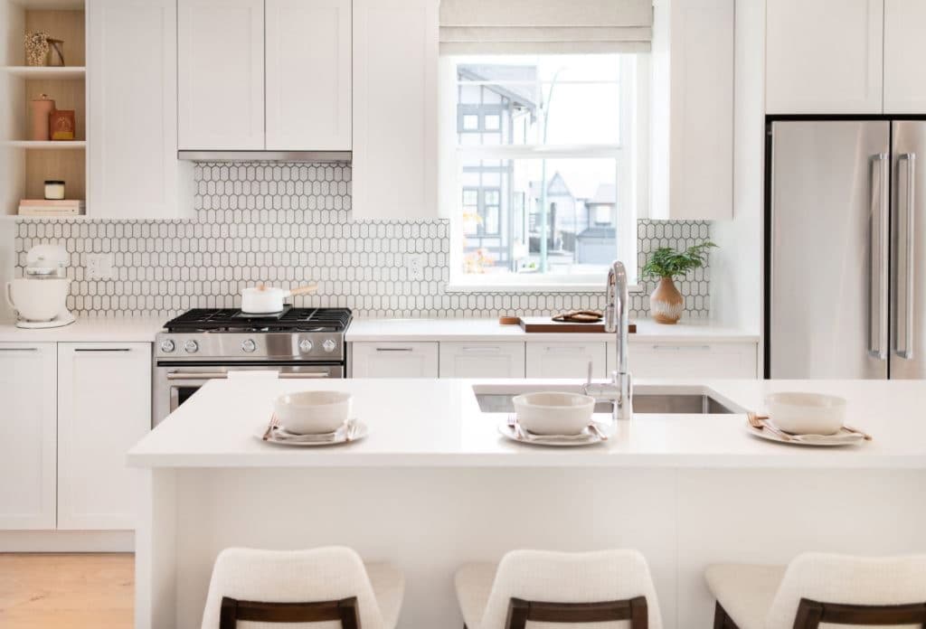 Baycrest West Condos – White kitchen with hexagon tile backsplash, stainless fridge and range, and an island set with bowls and plates for three.