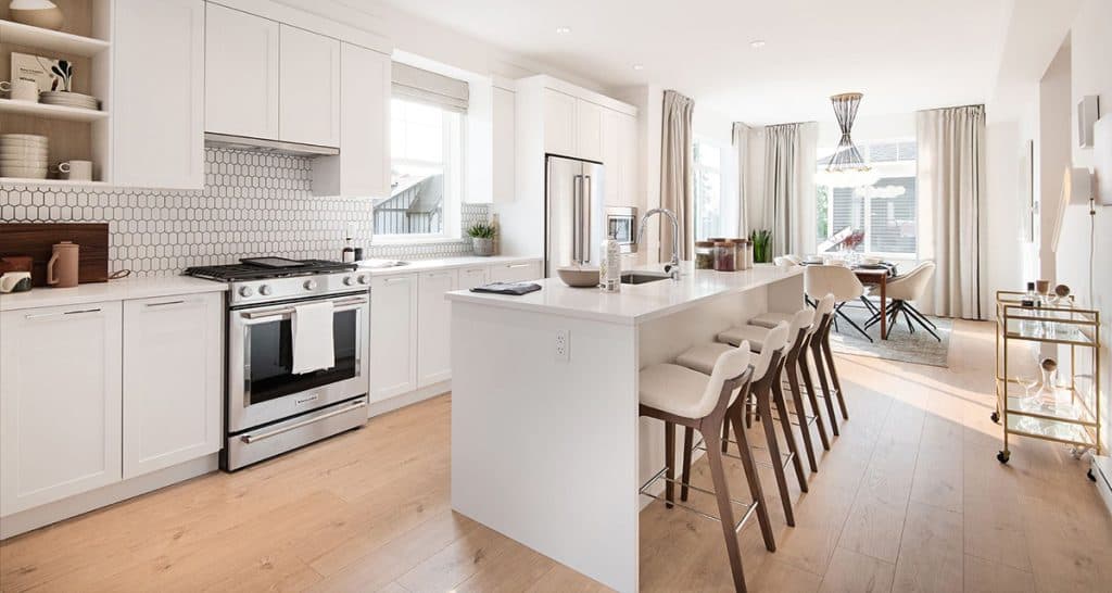 Baycrest West Condos – Open-concept white kitchen with a hexagon tile backsplash, stainless range, long island with bar stools, and a dining area beyond with a bar cart.