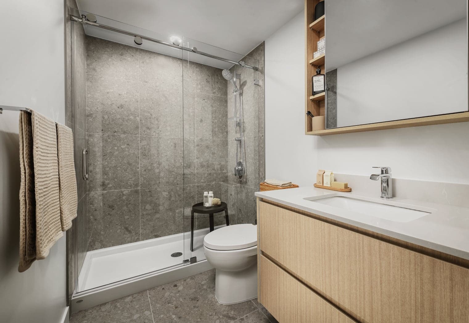 Gardena – A bathroom with a glass-enclosed tiled shower with a rain head, a toilet, and a wood vanity with a built-in shelving mirror cabinet.