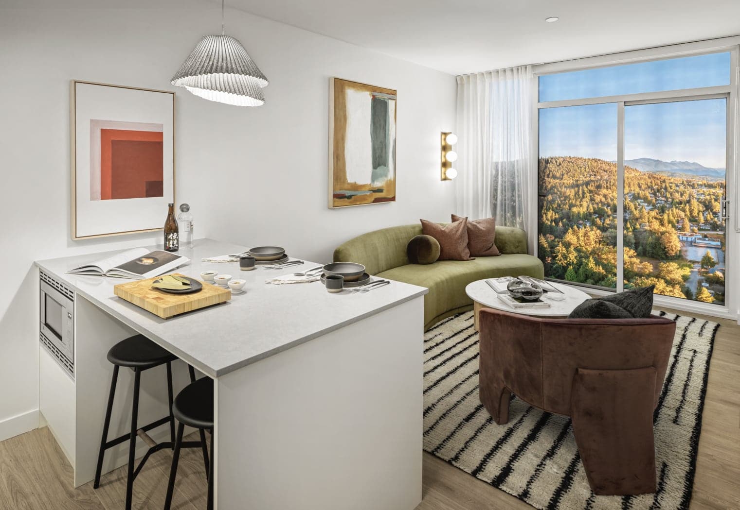 Gardena – An open kitchen and living space with a white waterfall-edge island set for dining, a curved olive green sofa, and a floor-to-ceiling window looking over forested hills.