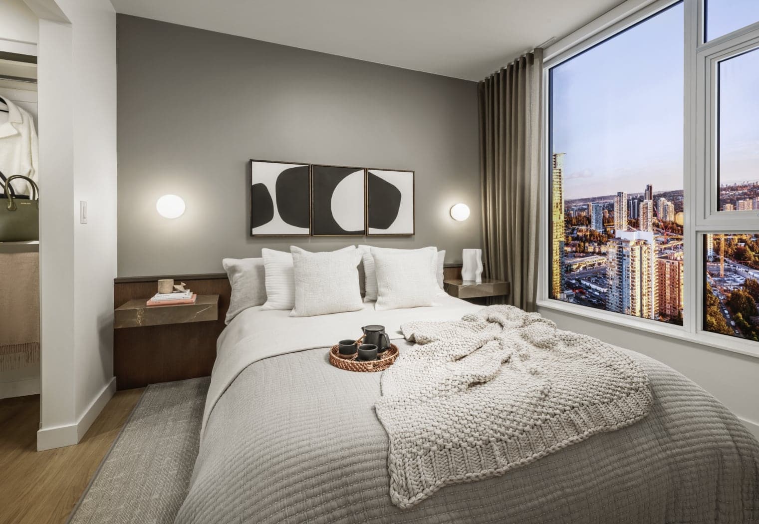 Gardena – A bedroom with a made bed, triptych abstract art above the headboard, wall sconces, and a large window with a high-rise city view at dusk.