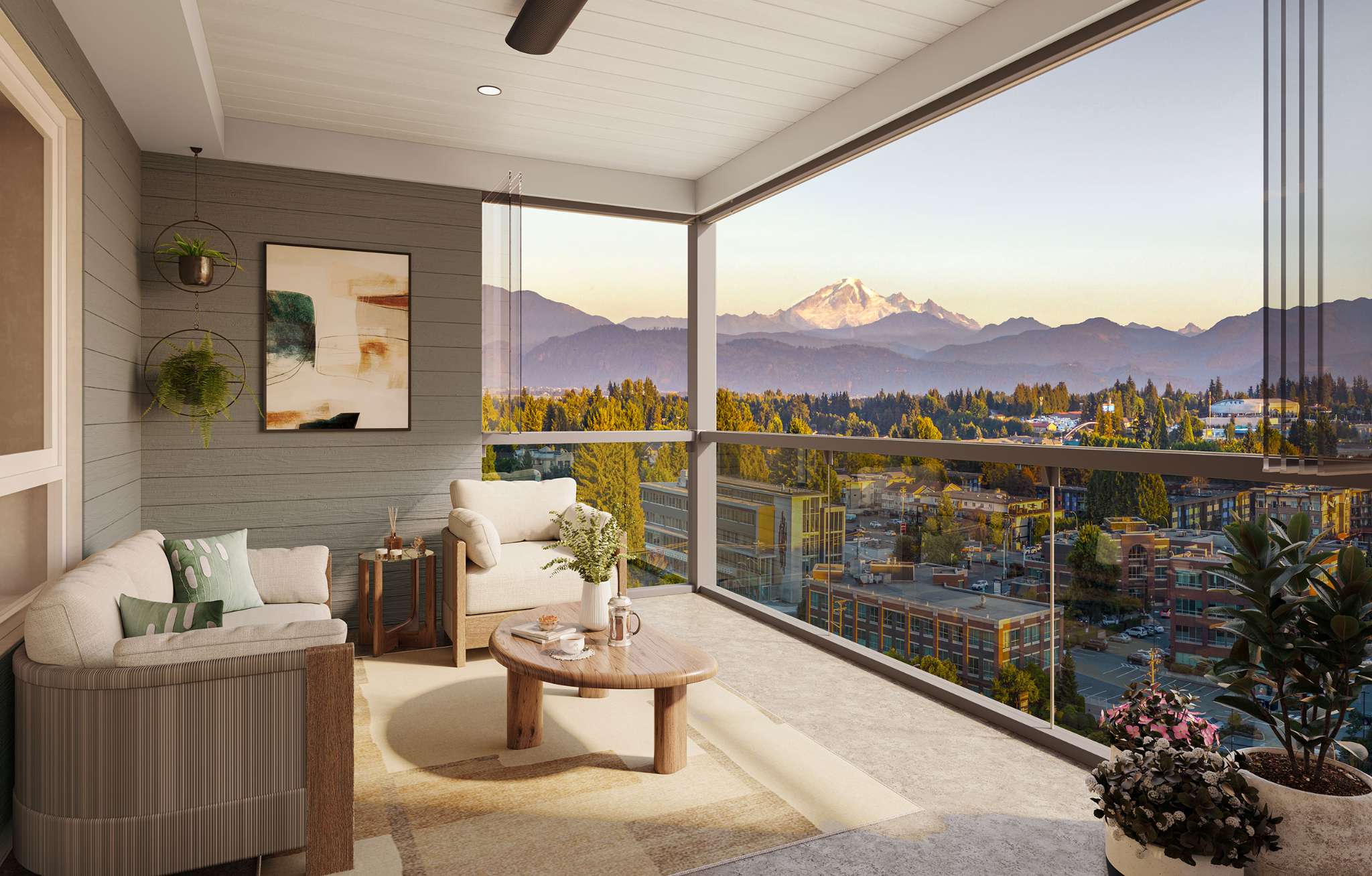 Southbend – A covered corner balcony furnished with a modern loveseat, armchair, and coffee table overlooks a panoramic landscape of a low-rise town, lush evergreen forests, and a prominent snow-capped mountain peak in the distance. Clear glass railings and retractable window panels frame the bright, golden-hour scenic vista.