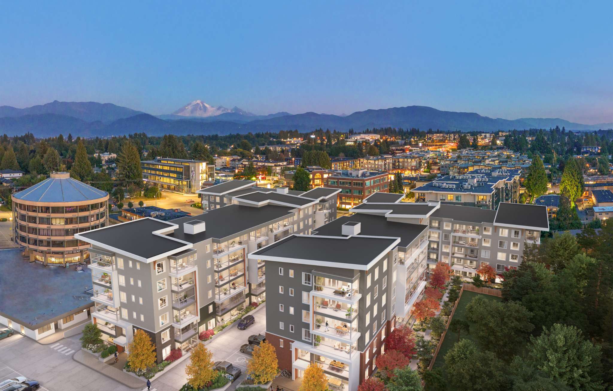 Southbend – An elevated twilight rendering of a multi-building mid-rise residential complex surrounded by a sprawling town, dense greenery, and glowing streetscapes. In the background, a dramatic mountain range with a prominent snow-capped peak rises against a clear evening sky.