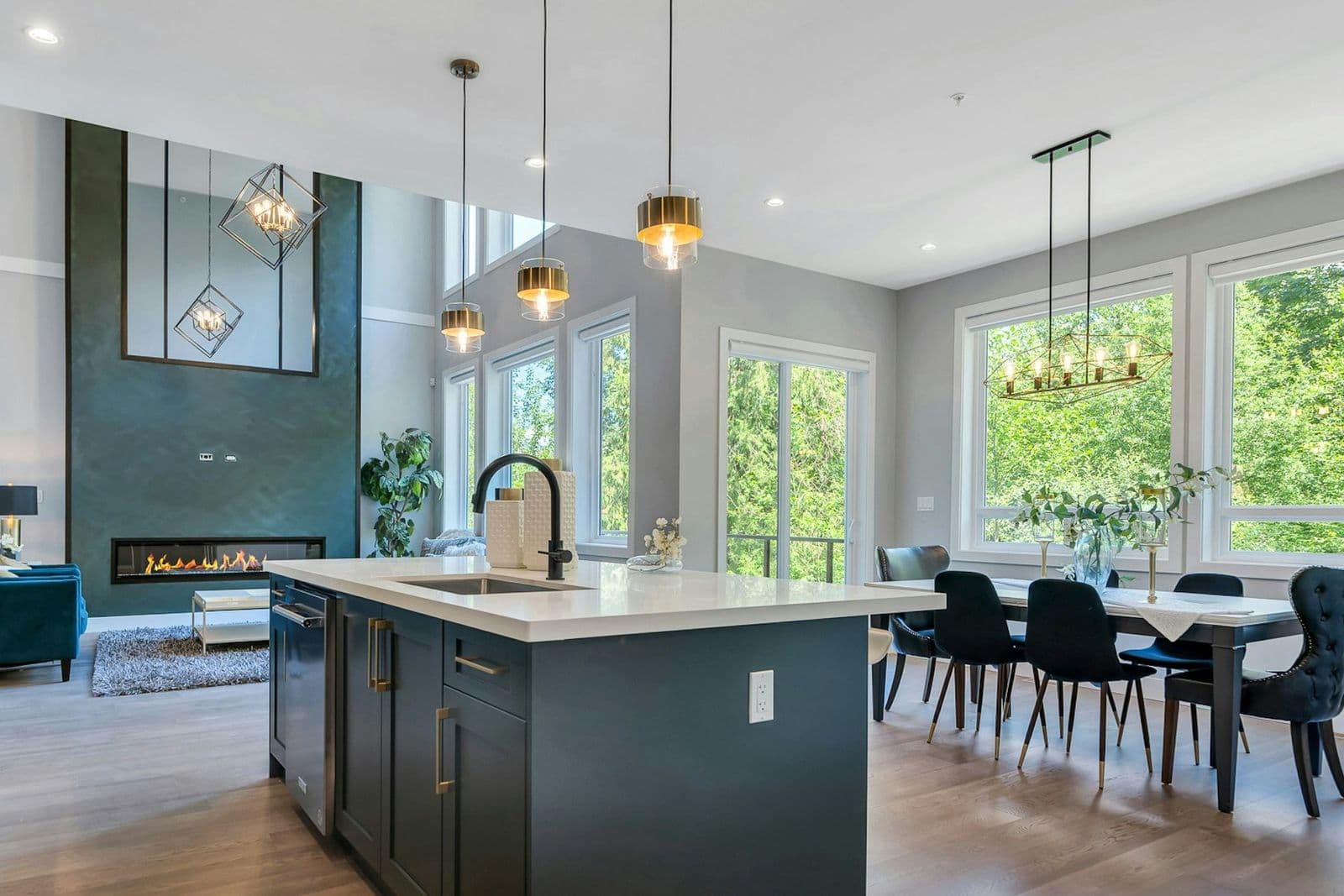 Marble Hill – Open-concept kitchen and dining room with a dark blue island, gold pendant lighting, a linear gas fireplace, and a dining table seating six near large windows.