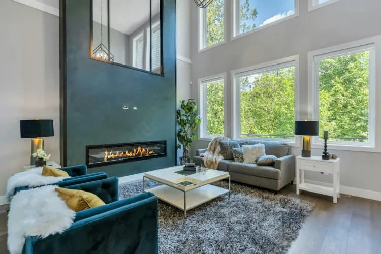 Marble Hill – Living room with a two-story fireplace feature wall, teal accent chairs, a grey sofa, shag rug, and large windows looking onto trees.