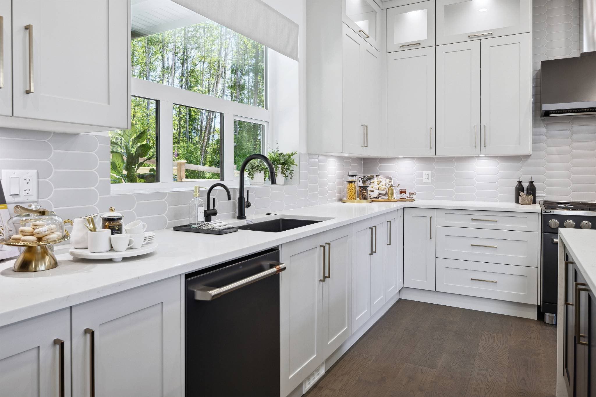 Timbers Edge – Second kitchen view with white shaker cabinets, black matte faucet and dishwasher, gold hardware, and a large window looking onto trees.