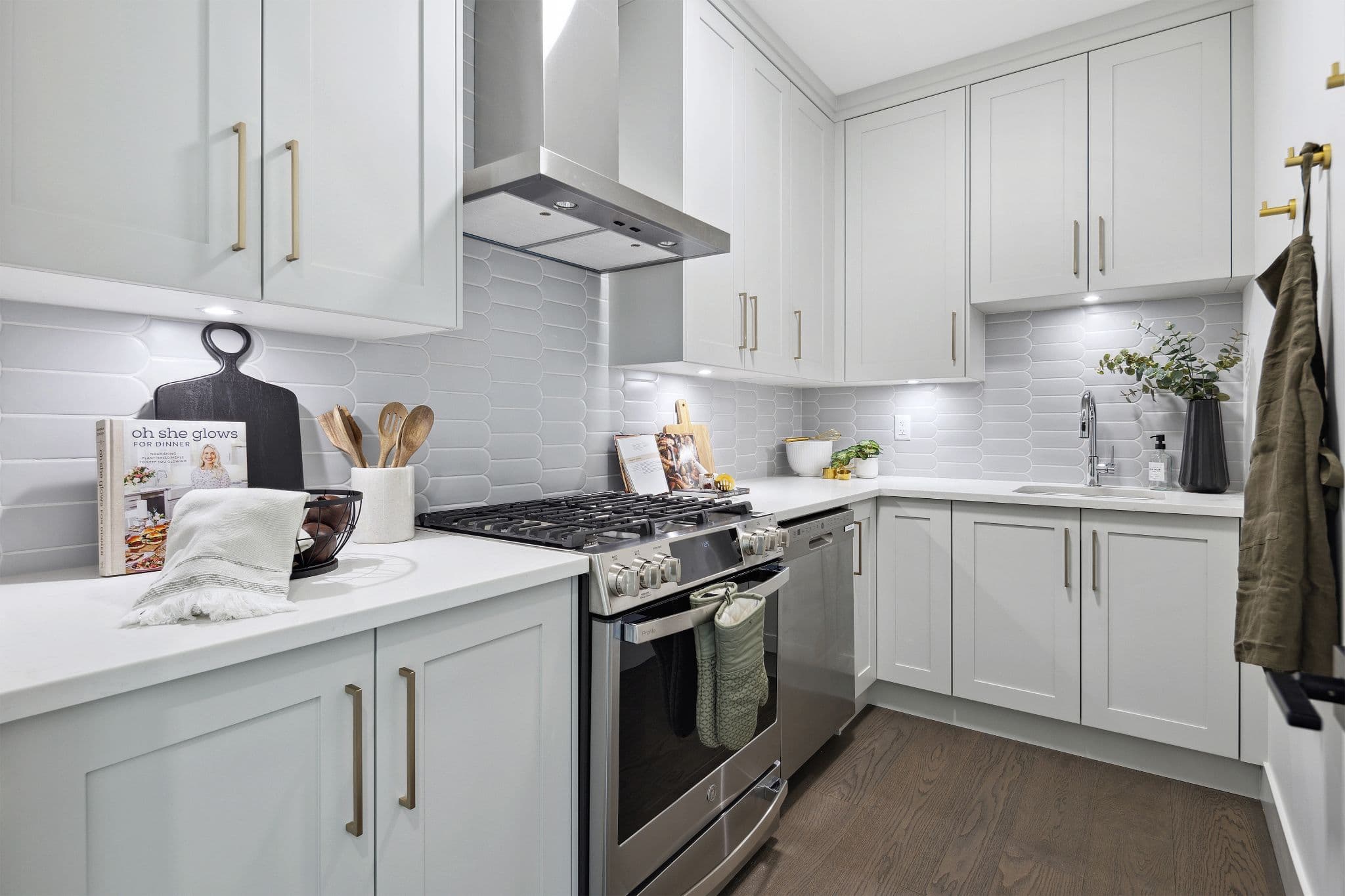 Timbers Edge – Kitchen with light gray shaker cabinets, gold hardware, patterned tile backsplash, stainless gas range and dishwasher, and dark wood flooring.