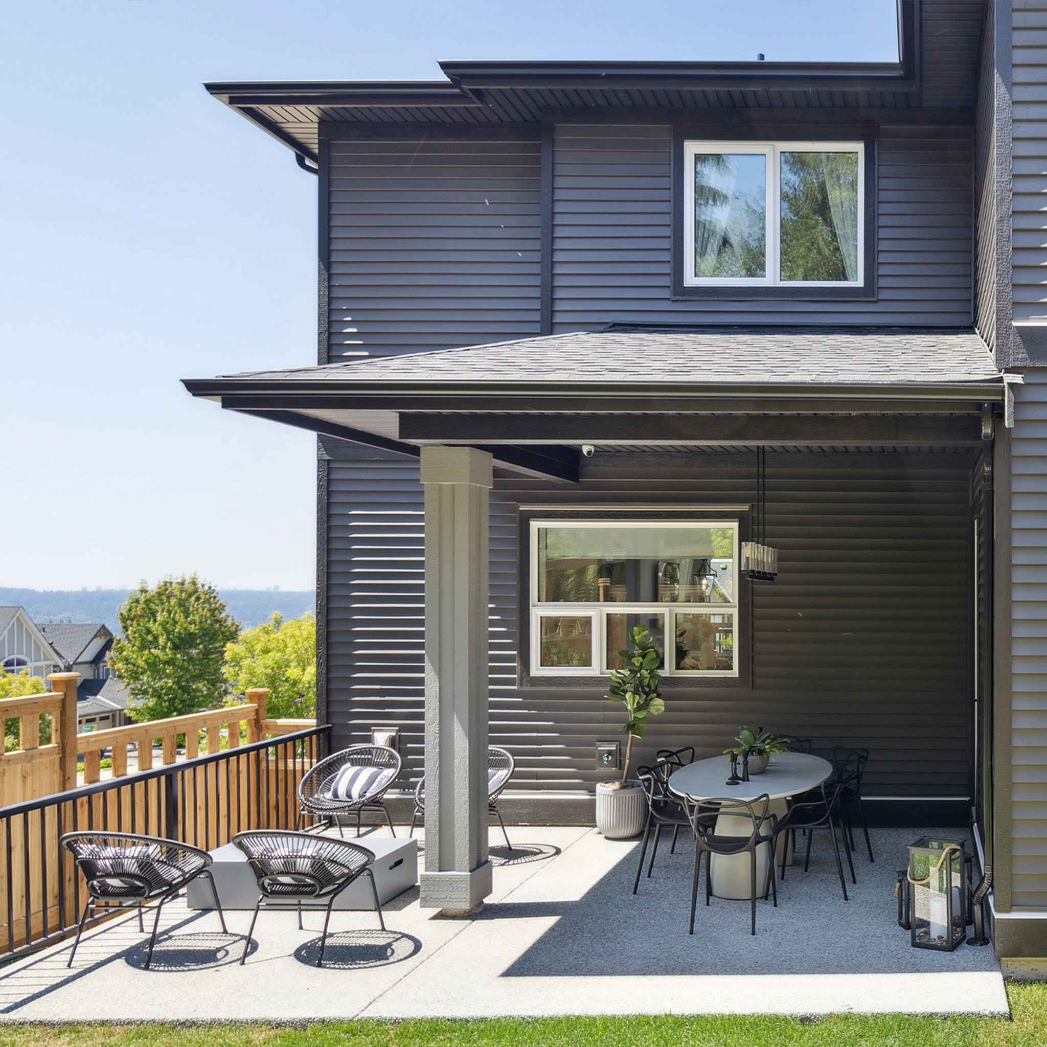 Timbers Edge – Covered back patio of the dark-sided house with outdoor dining table, lounge chairs, and a hillside neighborhood view in the distance.
