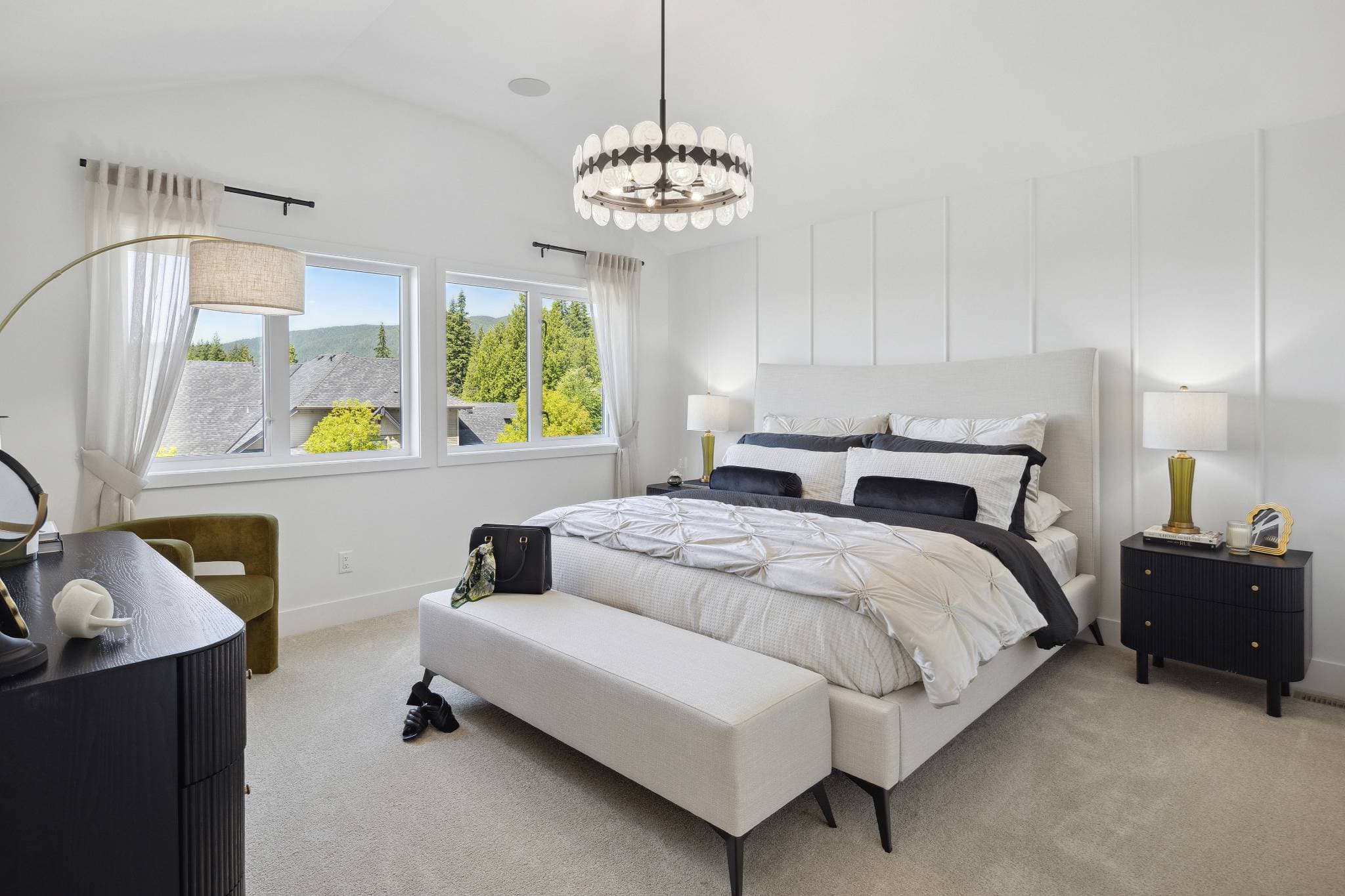 Timbers Edge – Primary bedroom with an upholstered light gray bed, dark nightstands, a round chandelier, and windows looking out to trees and mountains.