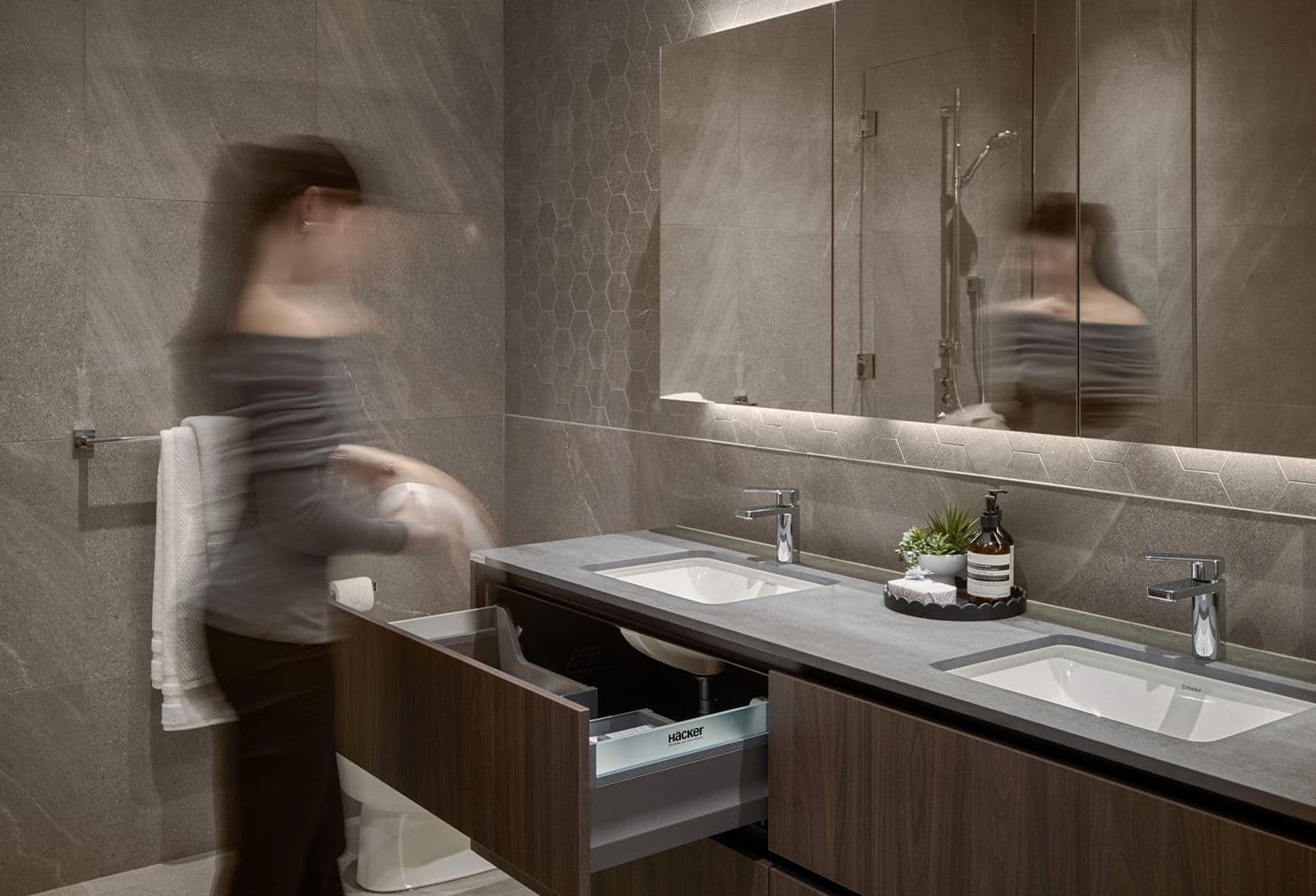 Fränk – A motion-blurred figure stands in a moody, modern bathroom featuring dark stone wall tiles, subtle hexagonal tile accents, and a large illuminated mirror cabinet. The space includes a floating dark wood vanity with dual undermount sinks, gray stone countertops, and a modern chrome faucet set.
