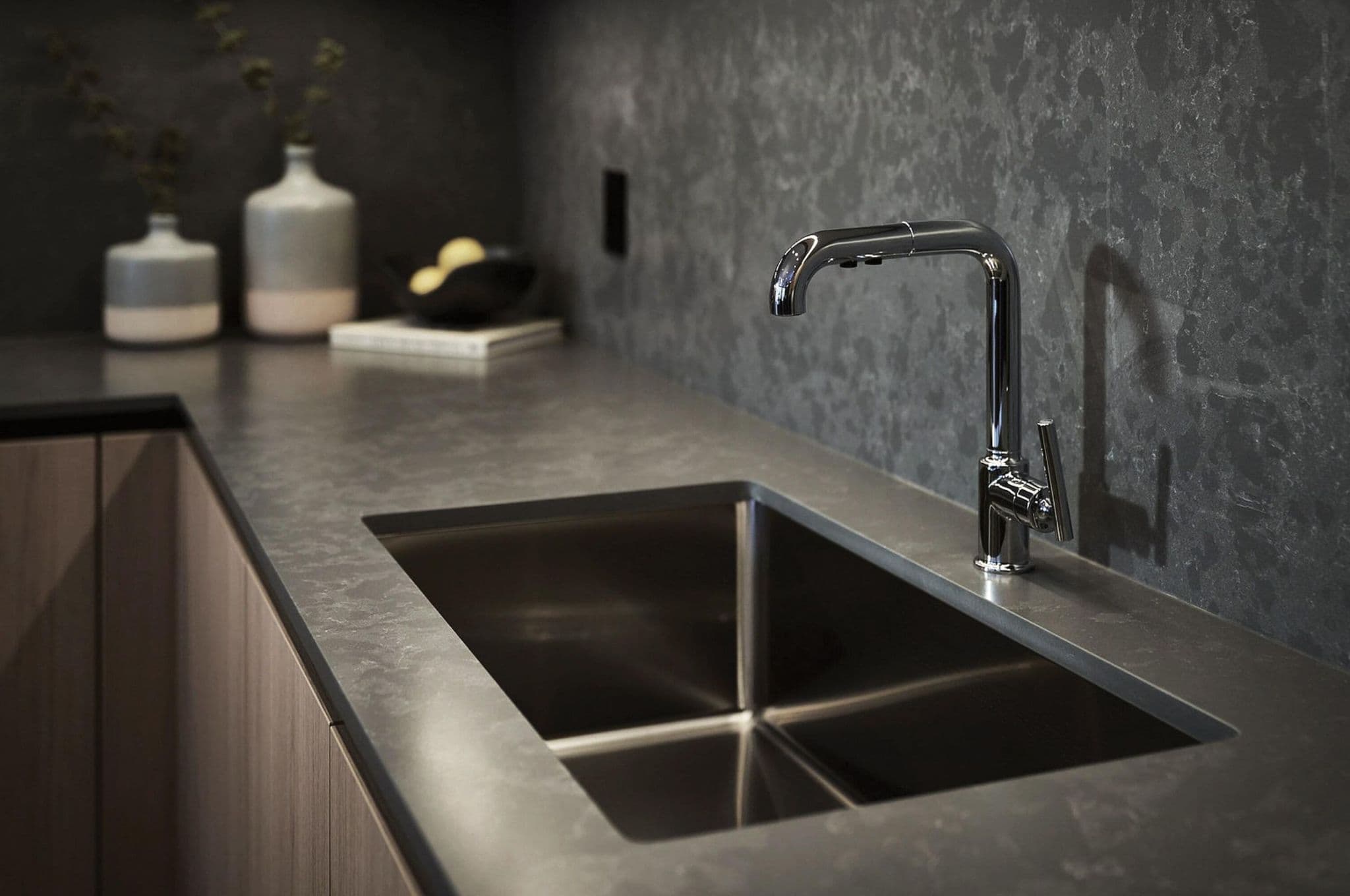 Fränk – A close-up shot of a modern kitchen featuring a polished chrome faucet and an undermount stainless steel sink set into dark, textured stone countertops. The background displays matching dark stone backsplashes and light wood lower cabinetry, complemented by subtle decorative ceramic vases for a sophisticated, moody feel.