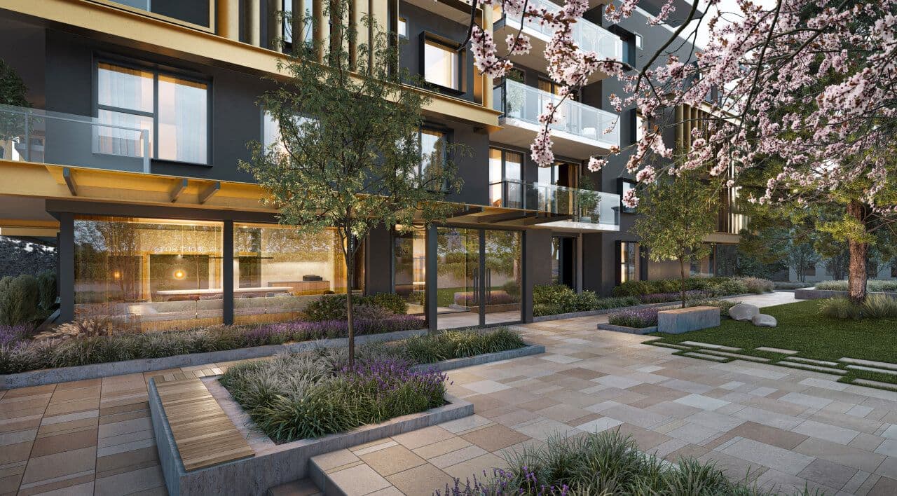 Fränk – Ground-level view of a modern residential building featuring dark grey siding, warm gold accents, glass balconies, and an illuminated lobby entrance. The paved courtyard is surrounded by stone planters filled with purple lavender, young trees, a bench, and a blooming cherry blossom tree in soft natural daylight.