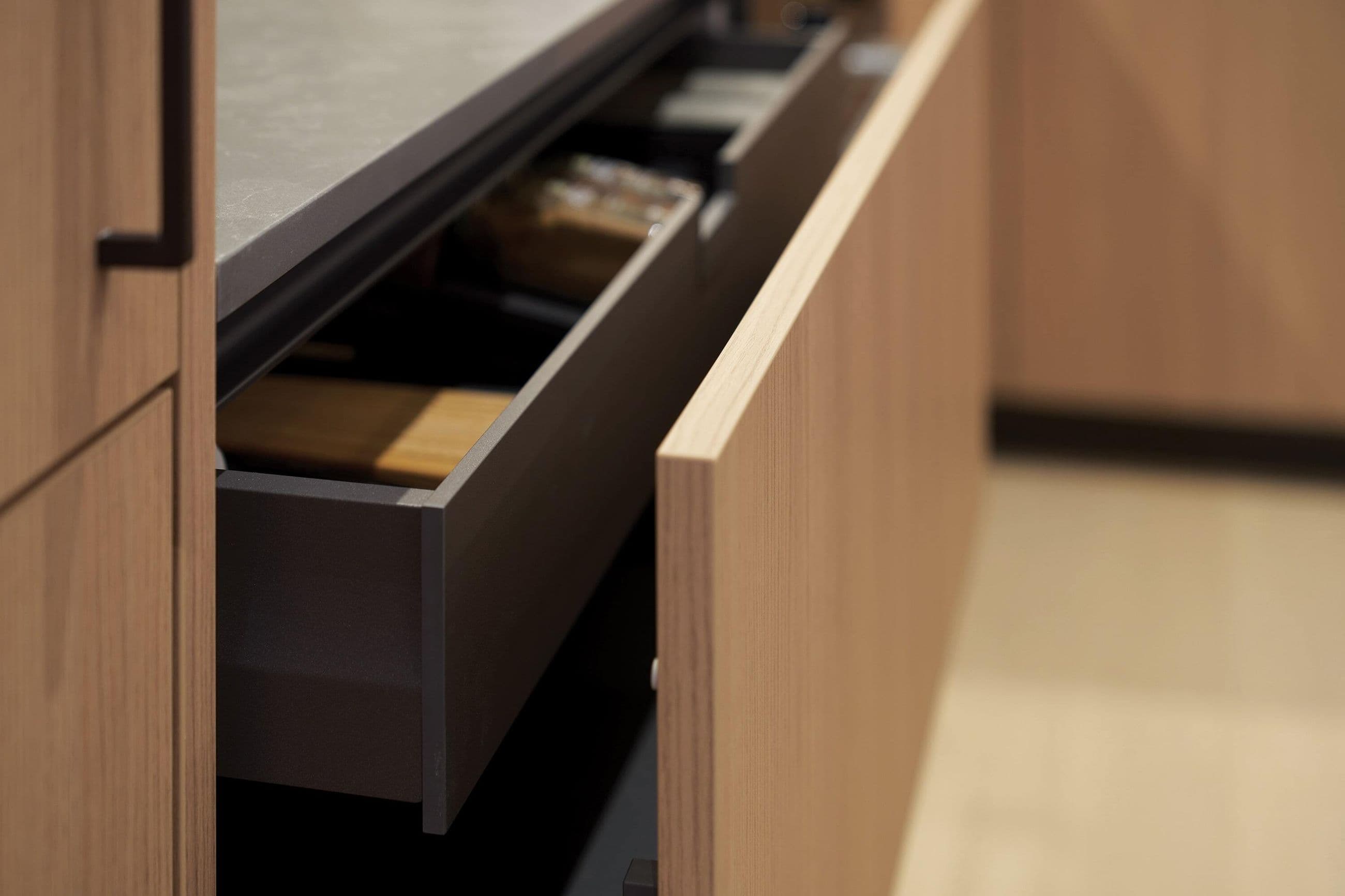 Fränk – A close-up, detail shot shows an open kitchen drawer featuring light wood grain paneling and a dark metal interior frame beneath a stone countertop. The soft, shallow depth of field highlights the craftsmanship and modern storage finishes of the cabinetry.