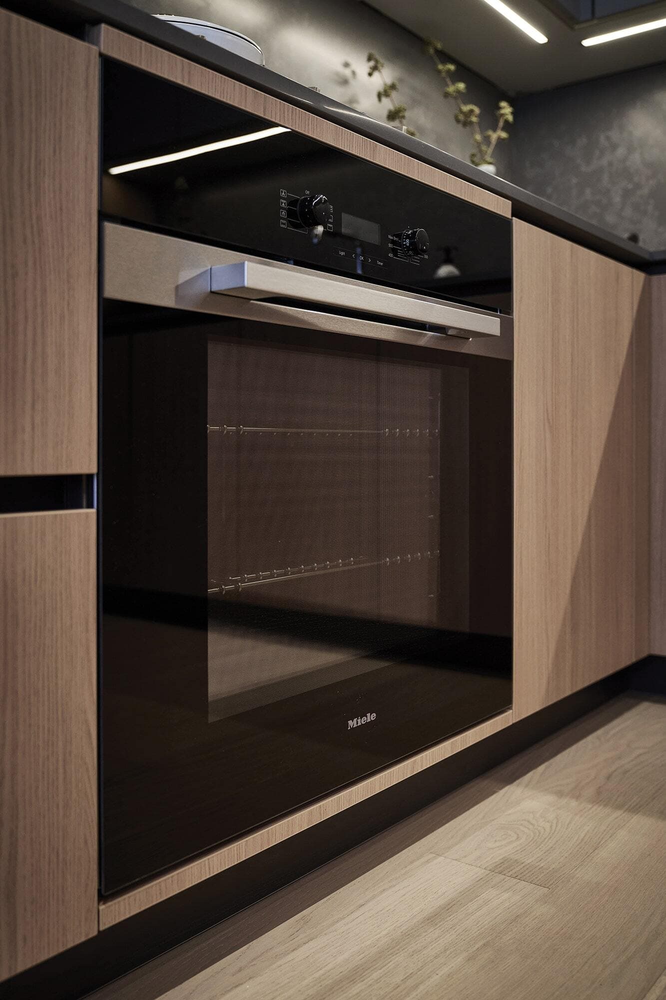Fränk – A close-up view of a sleek, black integrated Miele wall oven surrounded by flat-panel light wood cabinetry and dark countertops. Bright LED strip lighting glows along the ceiling edge, creating a modern, high-end kitchen aesthetic over light wood flooring.
