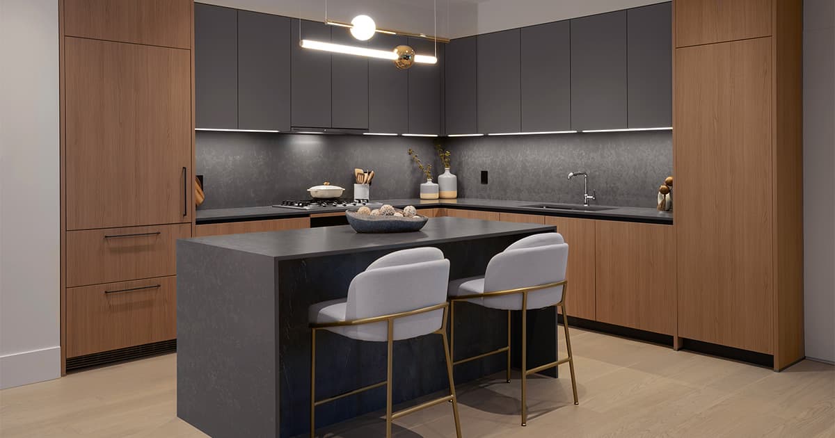 Fränk – A modern kitchen featuring matte charcoal upper cabinets, warm wood-toned lower cabinets, dark stone countertops, and a matching full-height backsplash with integrated under-cabinet lighting. A waterfall kitchen island in dark stone sits in the foreground with two upholstered barstools framed in brass hardware beneath a minimalist contemporary pendant light.