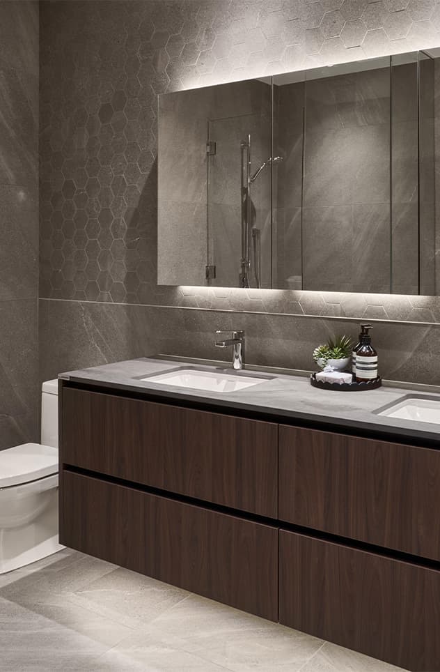 Fränk – A modern bathroom featuring a floating dark wood dual-sink vanity with a grey stone countertop, sleek chrome fixtures, and a large mirrored medicine cabinet with soft integrated under-lighting. The walls and floors are finished in moody charcoal grey tile with a geometric honeycomb texture behind the mirror, creating a luxurious and sophisticated atmosphere.