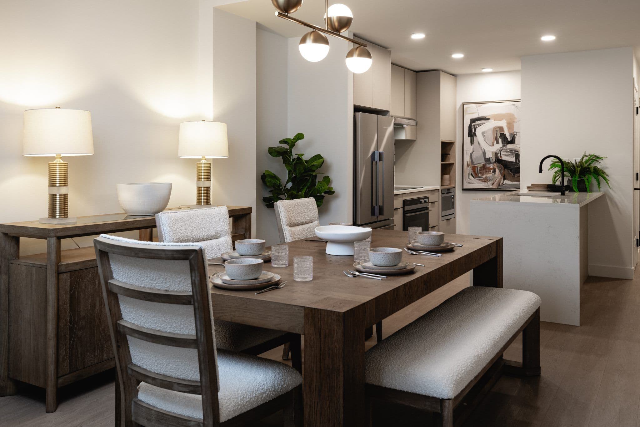Innova – Dining area with a wood table set for a meal, gold table lamps on a sideboard, and a kitchen with a stainless fridge in the background.