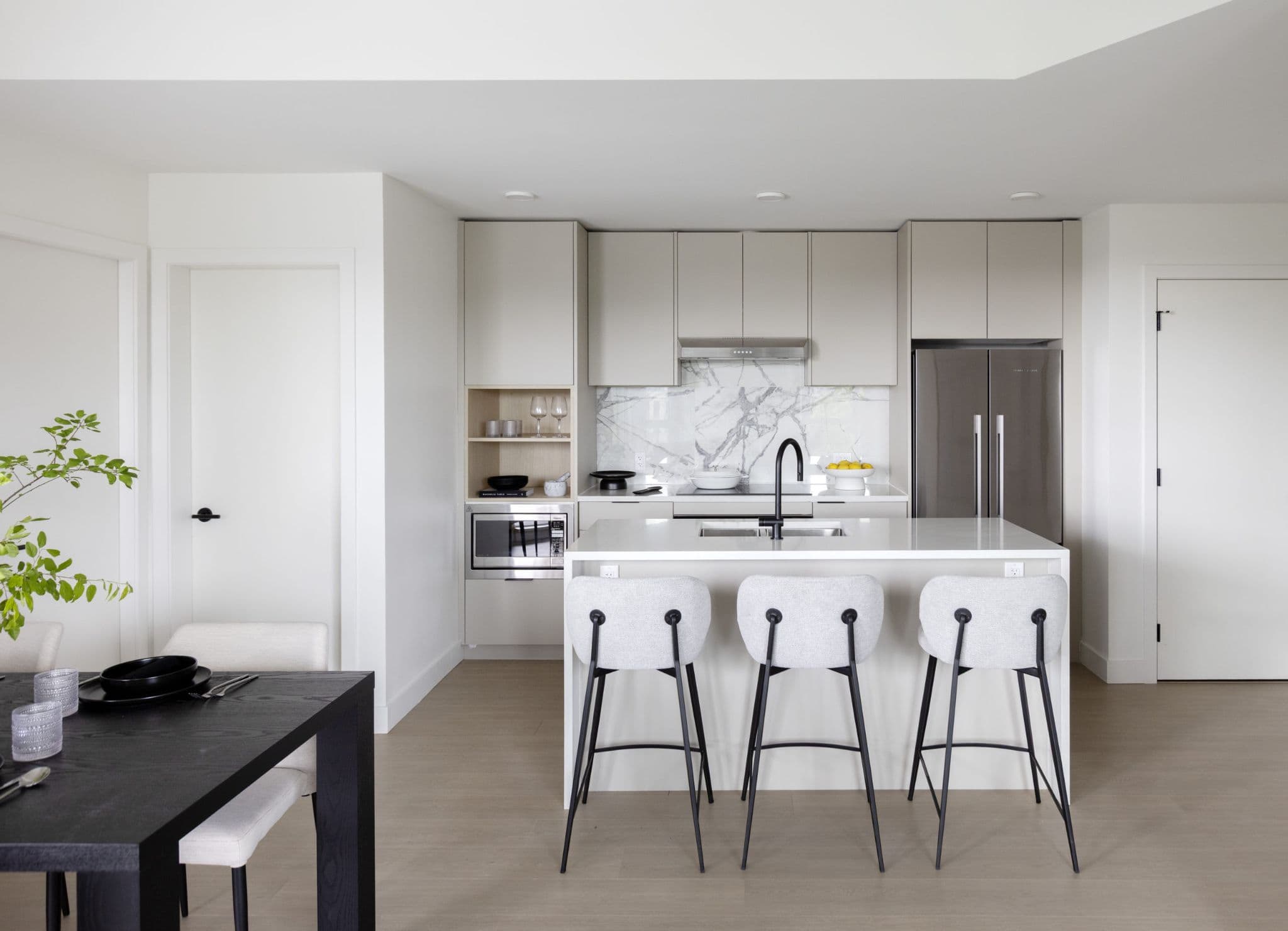 Innova – Kitchen with light wood cabinetry, a stainless fridge, and a white island with three bar stools, opening to a dining table.
