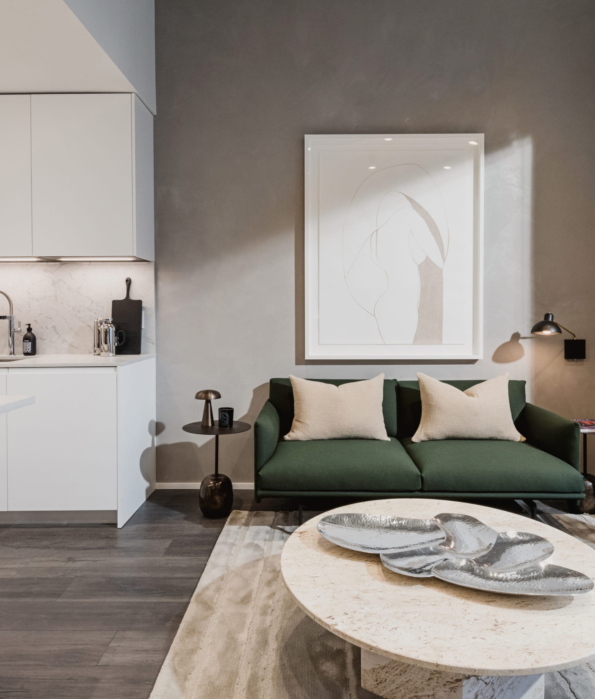 The Amazing Brentwood – Modern living area with a green sofa, cream throw pillows, round stone coffee table, and a white kitchenette visible to the side.