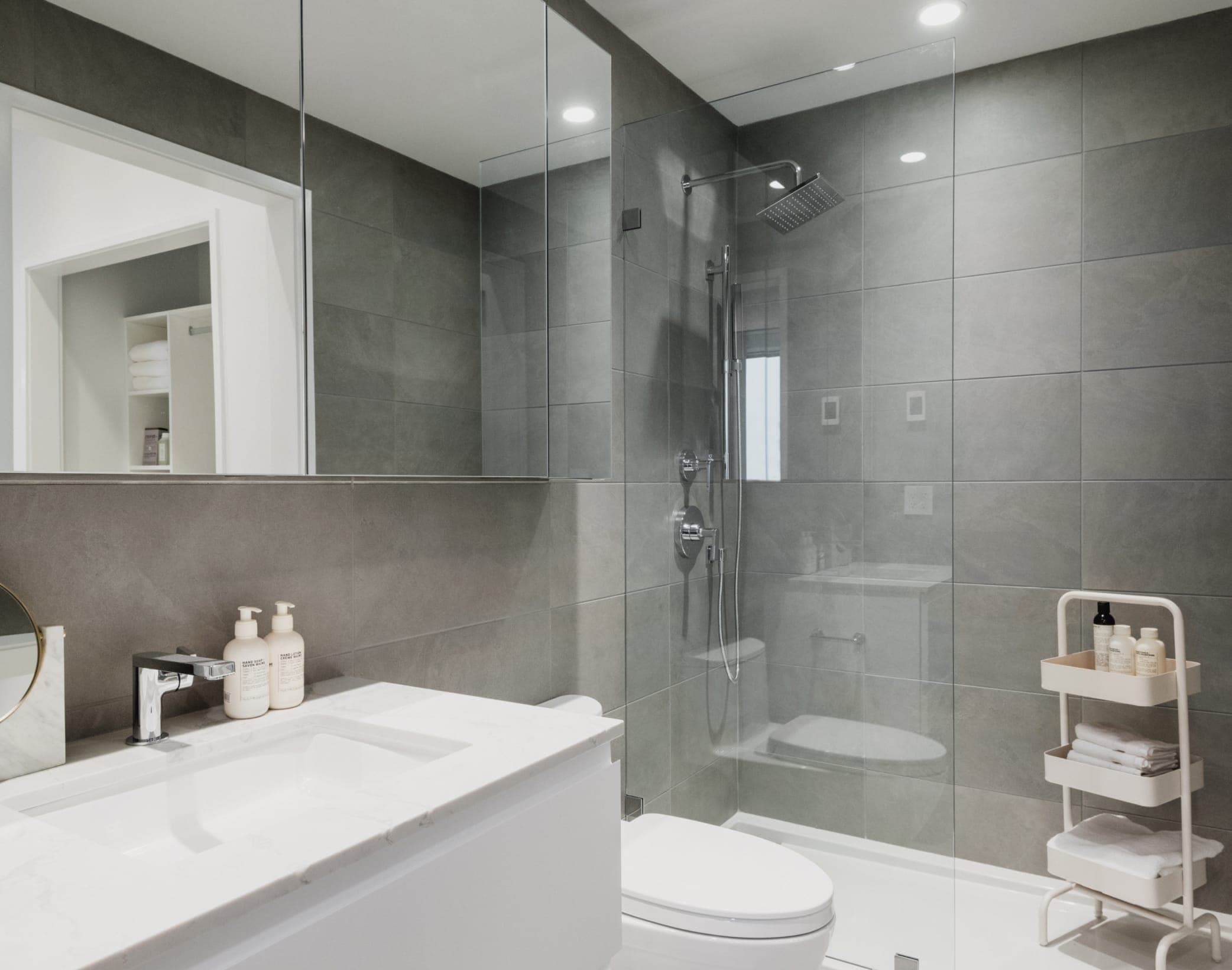 The Amazing Brentwood – Bathroom with a glass-enclosed shower, dark grey tile walls, a white vanity with sink, and a rolling bathroom caddy with towels.