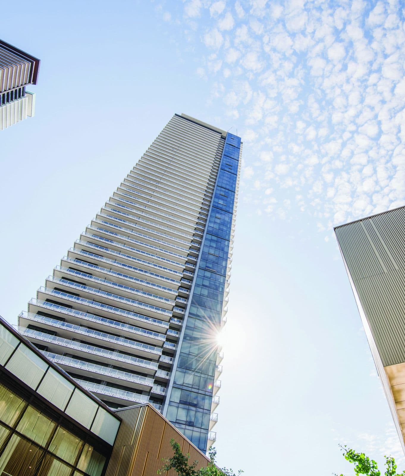 The Amazing Brentwood – Low upward-looking view of a tall glass and white balcony-clad residential tower against a blue sky with scattered clouds.