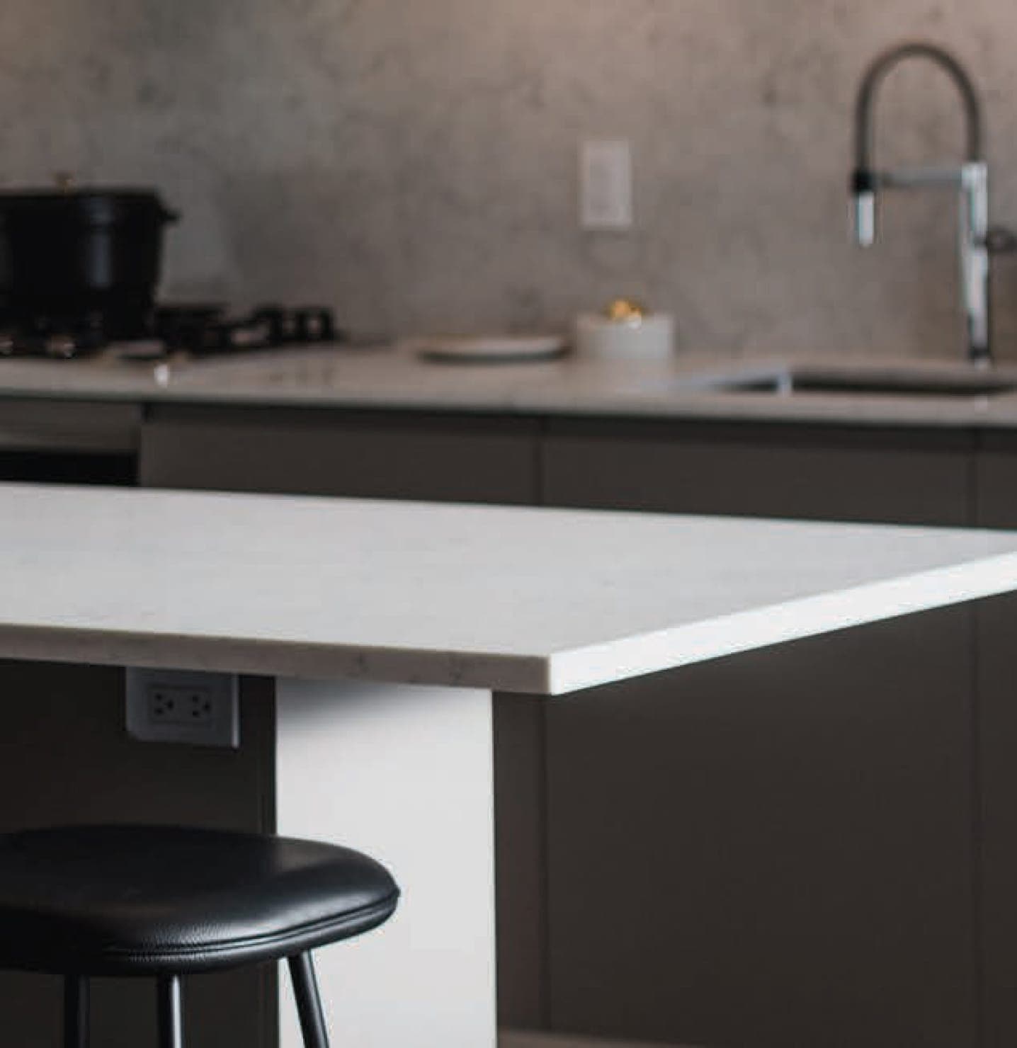 The Amazing Brentwood – Close-up of a kitchen island countertop with a black stovetop, sink, and faucet in the background, and a bar stool in the foreground.