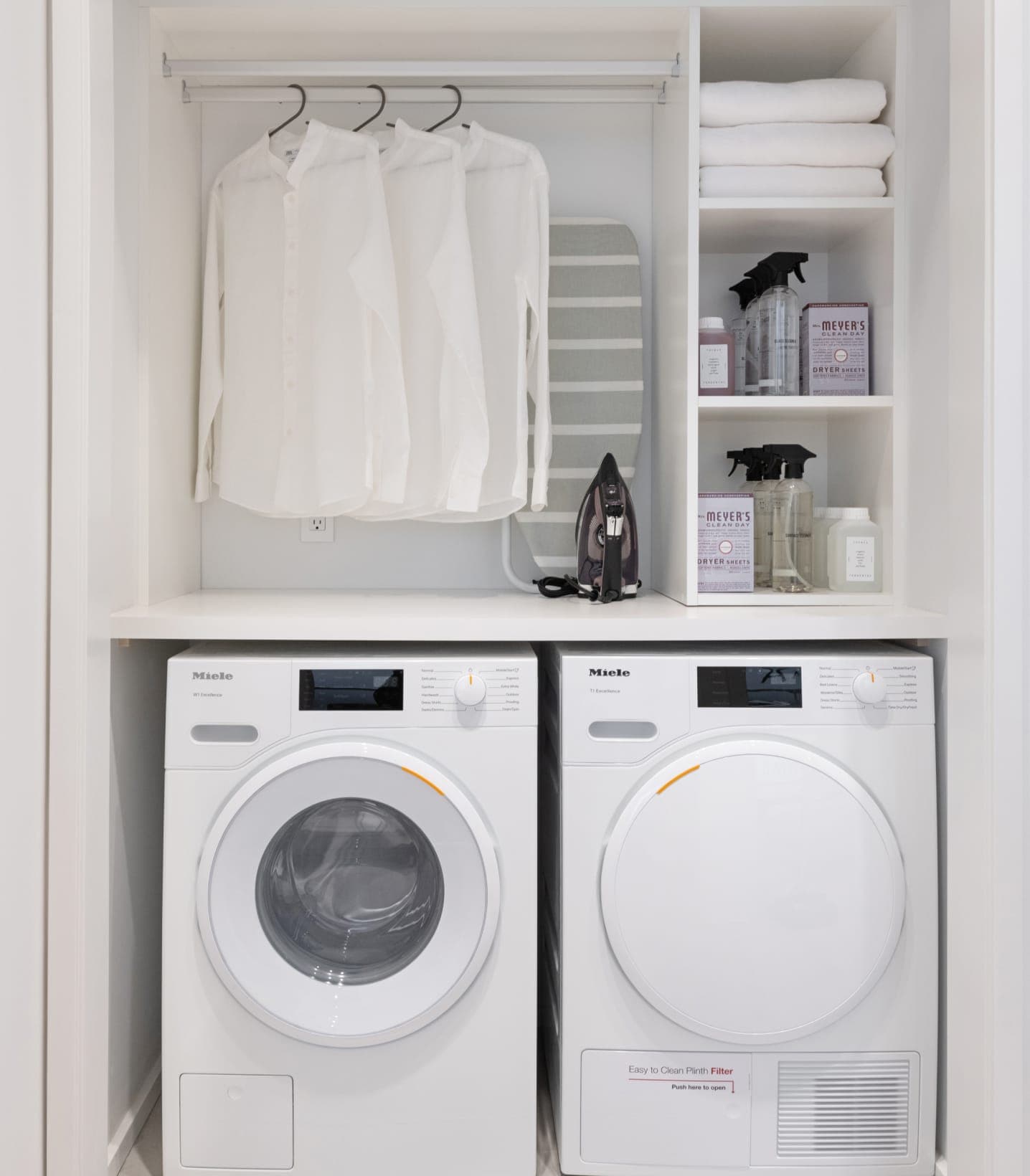 The Amazing Brentwood – Laundry closet with two white front-load Miele washer and dryer units, hanging shirts, an ironing board, and shelves of cleaning supplies.