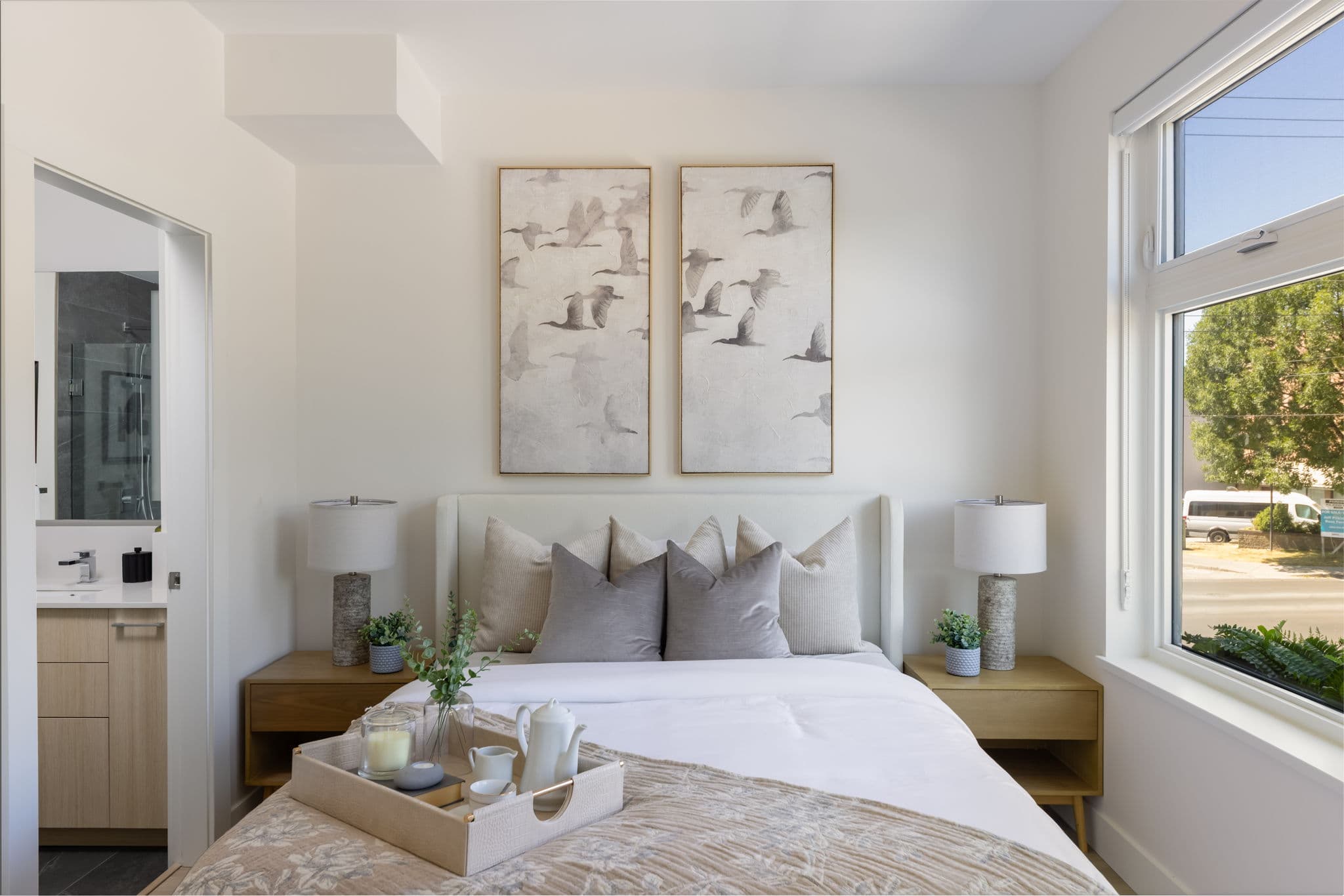 1515 Rupert – Bedroom with a white upholstered headboard, gray and cream pillows, two bird-print wall art panels, wood nightstands with lamps, and an ensuite bathroom visible through an open door.