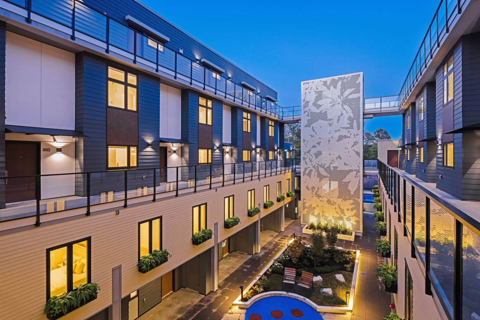1515 Rupert – Evening view of the building's interior courtyard with a decorative perforated art screen, garden beds, wooden benches and a glimpse of a pool in the distance.