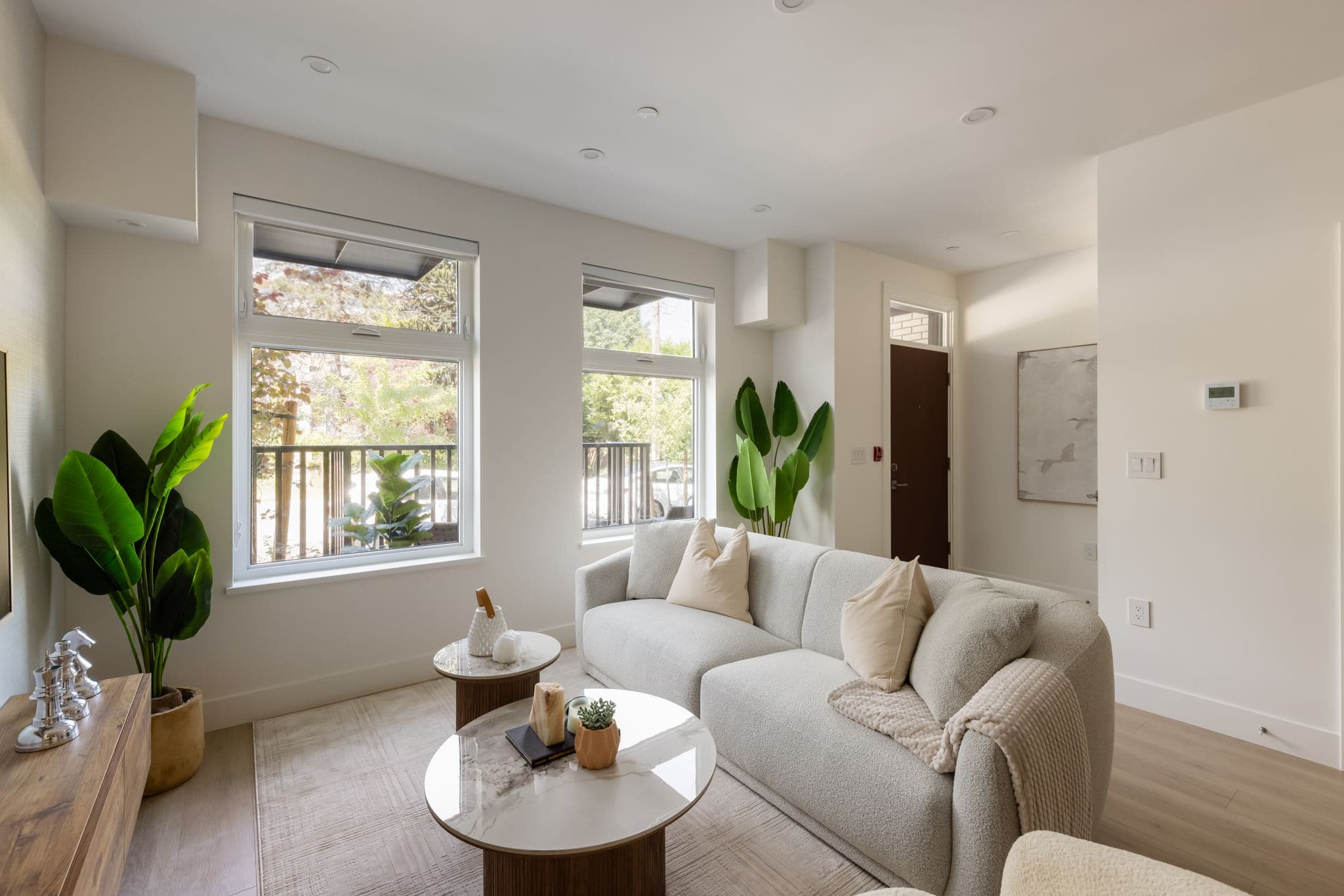 1515 Rupert – Bright living room with a light gray sofa, round marble coffee tables, a large potted plant, and two windows overlooking a balcony railing.