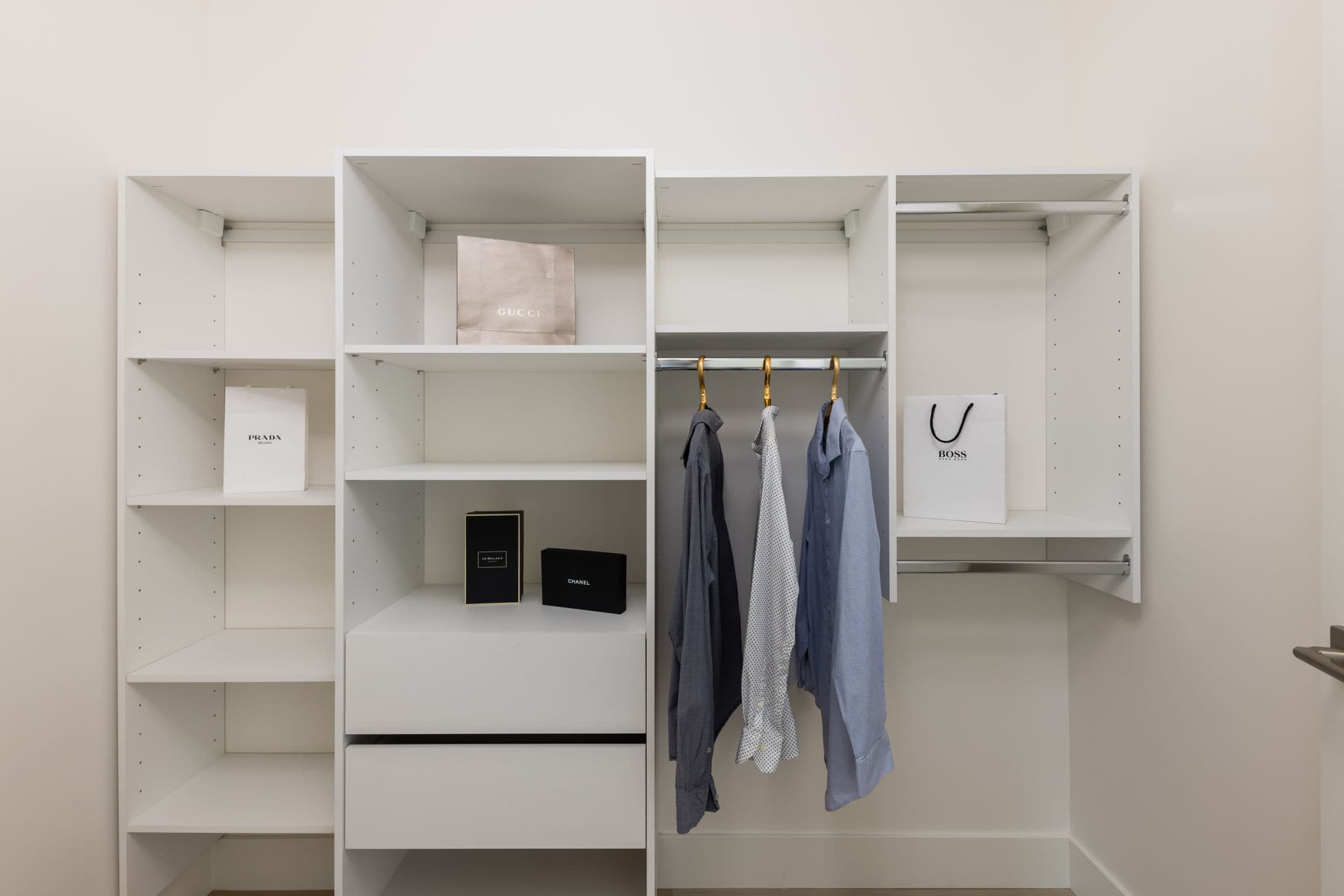 1515 Rupert – Walk-in closet with white built-in shelving, hanging rods with dress shirts, and empty designer shopping bags (Gucci, Prada, Boss) displayed as decor.