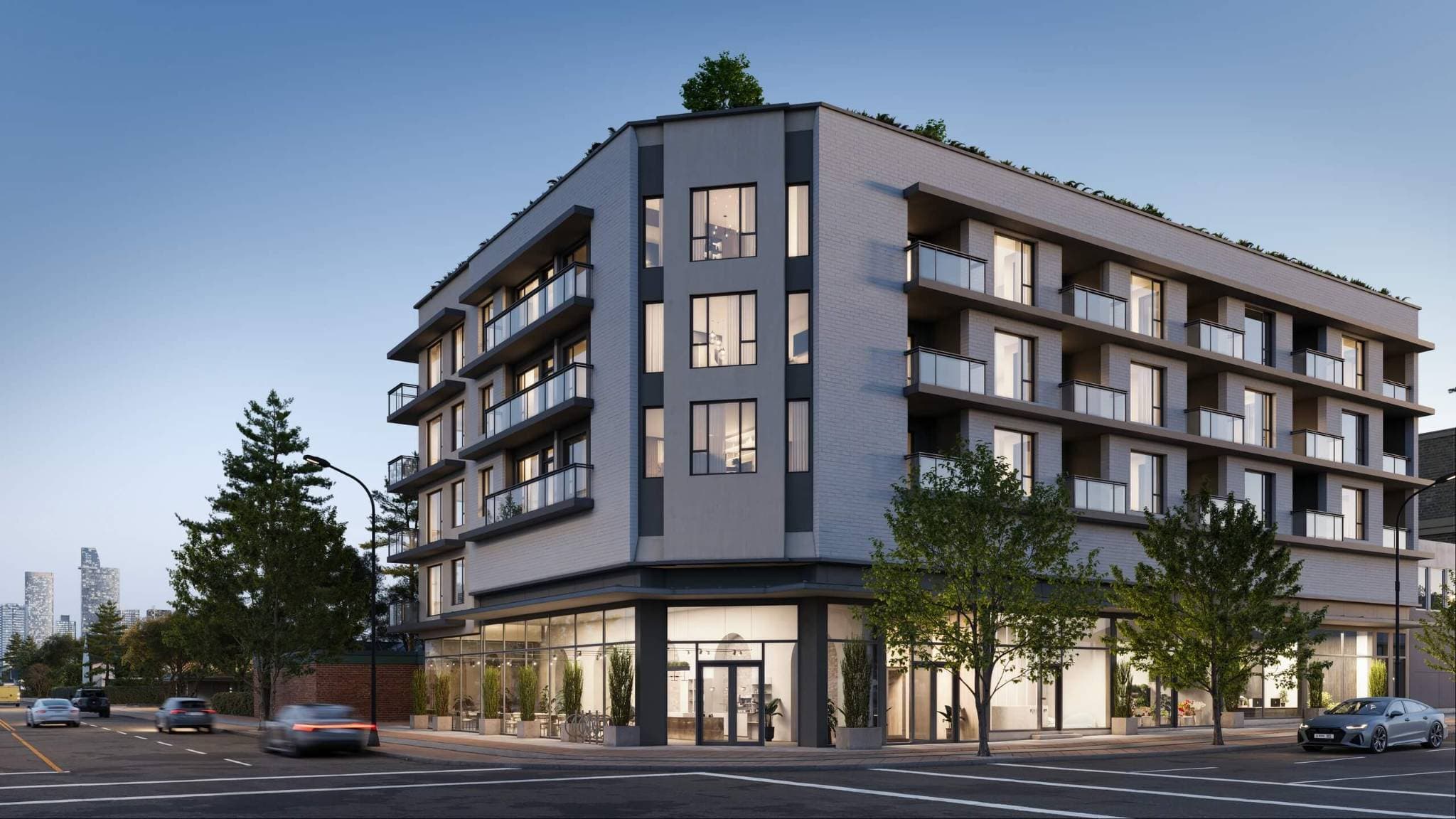 Centrale – A modern mid-rise residential building on a street corner featuring a light gray brick façade, glass-railed balconies, and illuminated floor-to-ceiling ground-level retail spaces. Cars drive along the adjacent asphalt street during dusk, with a distant city skyline visible under a clear, late-afternoon sky.