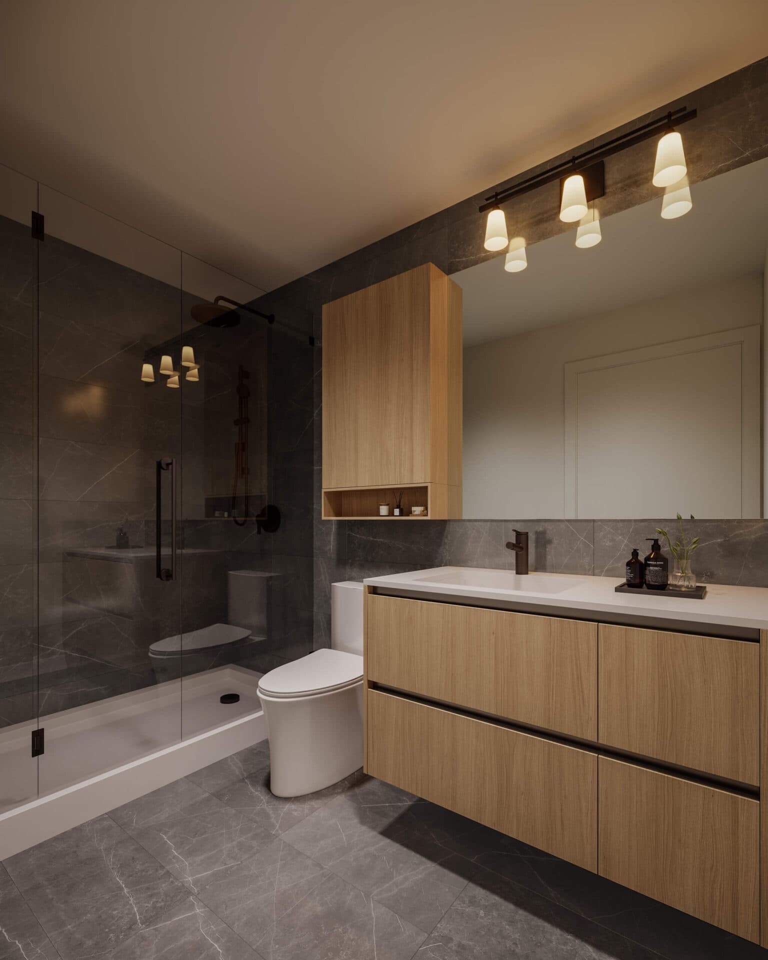 Centrale – A modern bathroom interior featuring dark gray stone wall and floor tiles paired with warm light wood cabinetry and vanity drawers. A frameless glass walk-in shower sits adjacent to the toilet and sink area, softly illuminated by overhead vanity lights.
